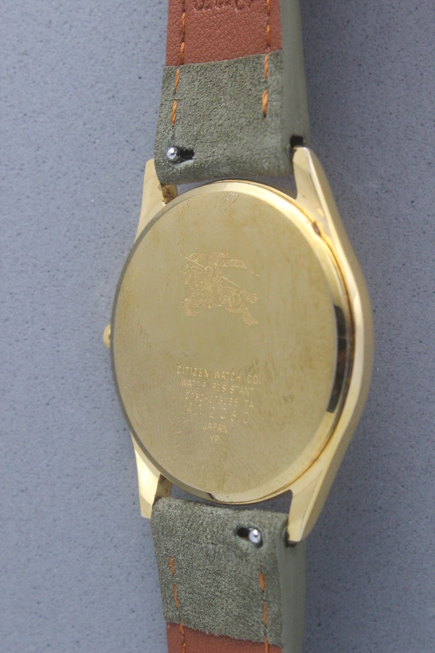 Back view of vintage Burberry wristwatch featuring a polished gold-tone caseback with engraved details, including the iconic Burberrys knight logo and inscriptions. The caseback specifies water resistance and model details: 2280-223465 TA, 412018 0, made in Japan. The watch is paired with a green suede strap and brown leather backing, showcasing aa blend of elegance and durability.