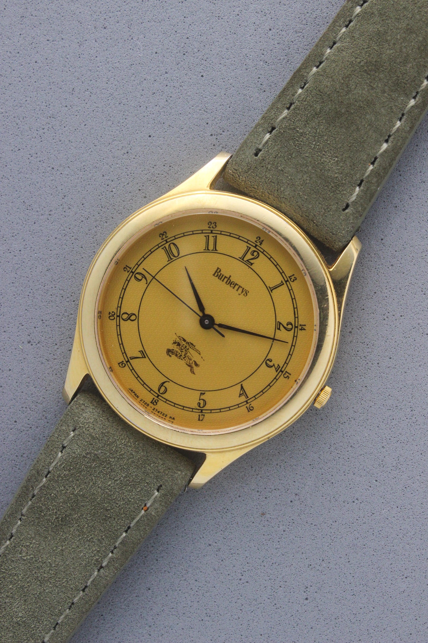 Vintage Burberrys wristwatch with a gold-tone round case, yellow dial featuring black Arabic numerals for both 12-hour and 24-hour markers, and an elegant black logo crest at the 6 o’clock position. The watch is paired with a green suede strap, showcasing a sophisticated and timeless design. Made in Japan, model number 2720-274722 KA.