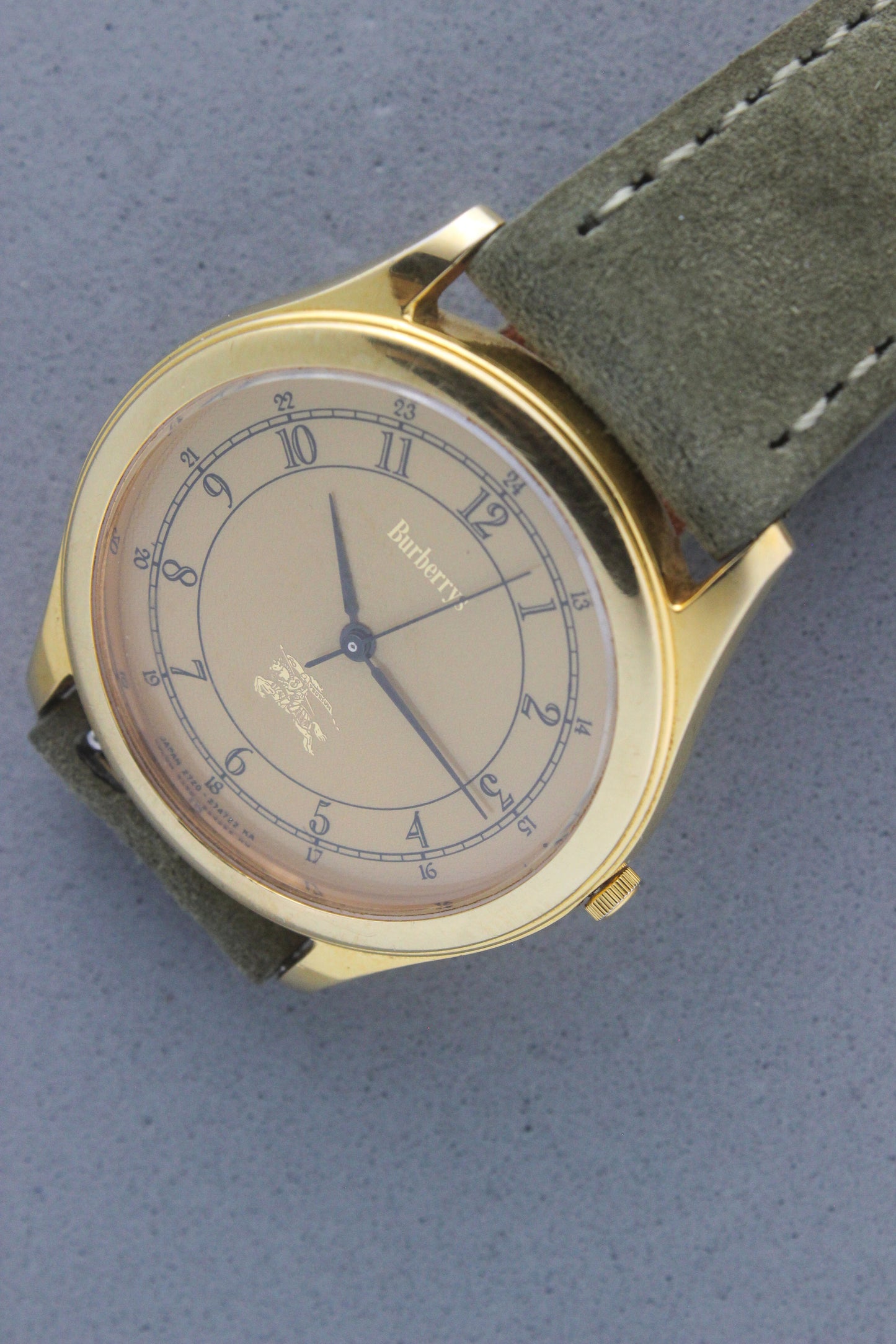 Vintage Burberrys wristwatch with a gold-tone round case, yellow dial featuring black Arabic numerals for both 12-hour and 24-hour markers, and an elegant black logo crest at the 6 o'clock position. The watch is paired with a green suede strap, showcasing a sophisticated and timeless design. Made in Japan, model number 2280-223465 TA, 412018 0,.