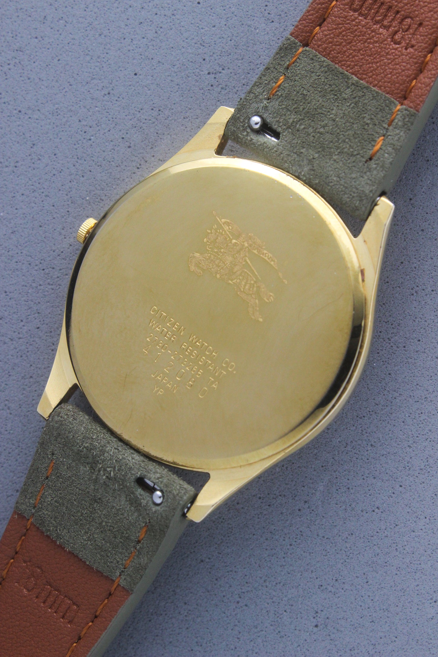 Back view of vintage Burberry wristwatch featuring a polished gold-tone caseback with engraved details, including the iconic Burberrys knight logo and inscriptions. The caseback specifies water resistance and model details: 2280-223465 TA, 412018 0, made in Japan. The watch is paired with a green suede strap and brown leather backing, showcasing aa blend of elegance and durability.