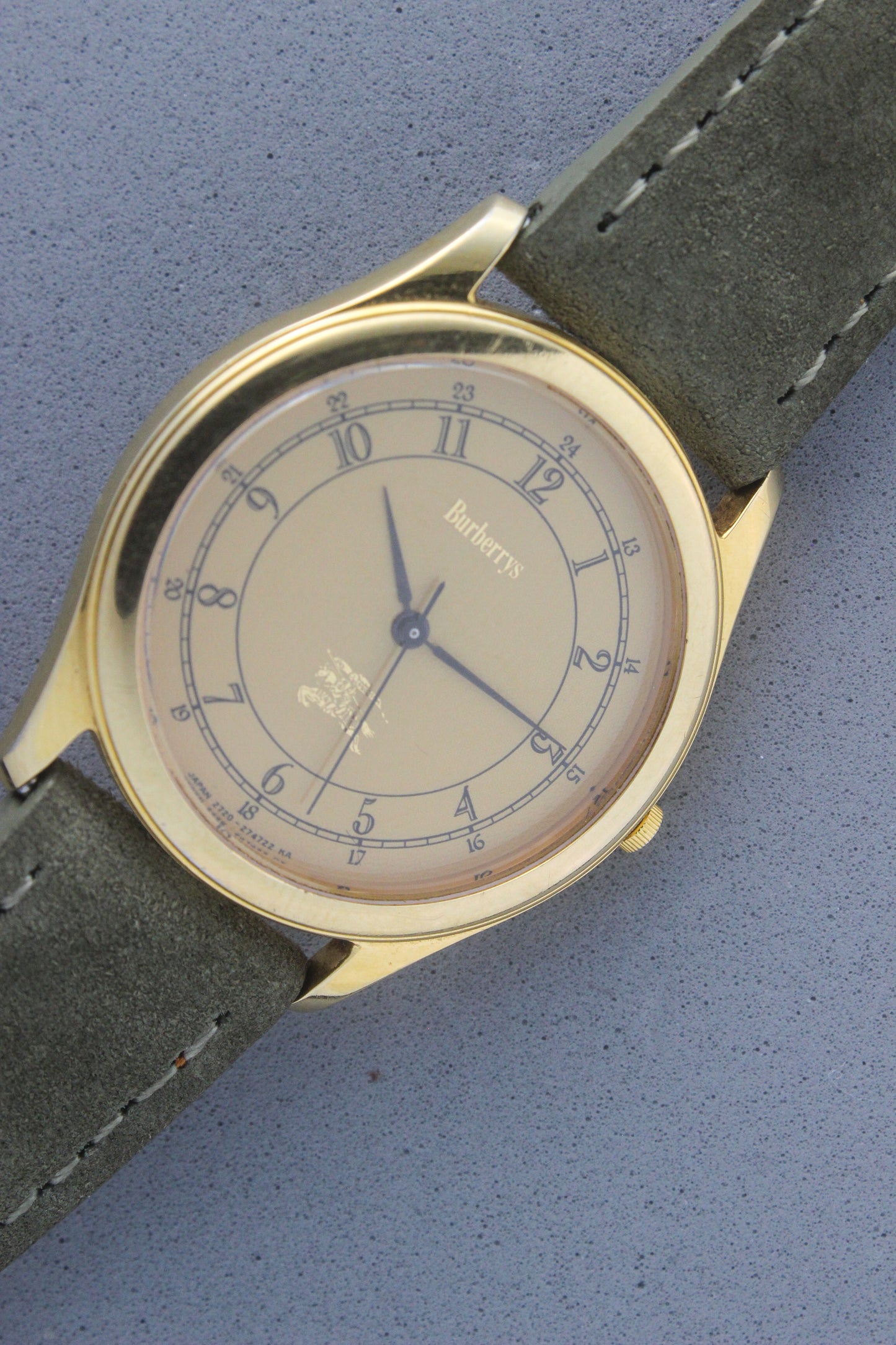 Vintage Burberrys wristwatch with a gold-tone round case, yellow dial featuring black Arabic numerals for both 12-hour and 24-hour markers, and an elegant black logo crest at the 6 o'clock position. The watch is paired with a green suede strap, showcasing a sophisticated and timeless design. Made in Japan, model number 2280-223465 TA, 412018 0.