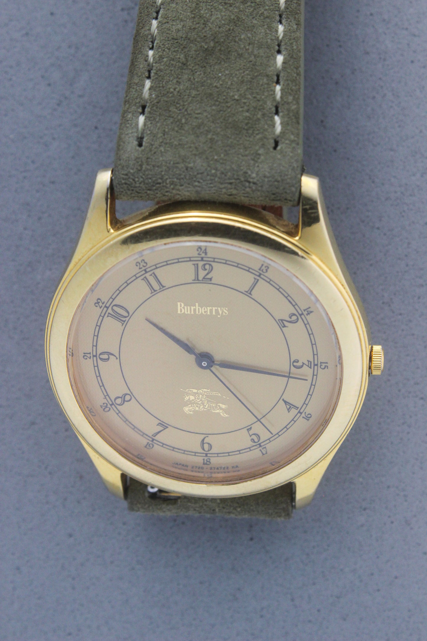 Vintage Burberrys wristwatch with a gold-tone round case, yellow dial featuring black Arabic numerals for both 12-hour and 24-hour markers, and an elegant black logo crest at the 6 o'clock position. The watch is paired with a green suede strap, showcasing a sophisticated and timeless design. Made in Japan, model number 2280-223465 TA, 412018 0.