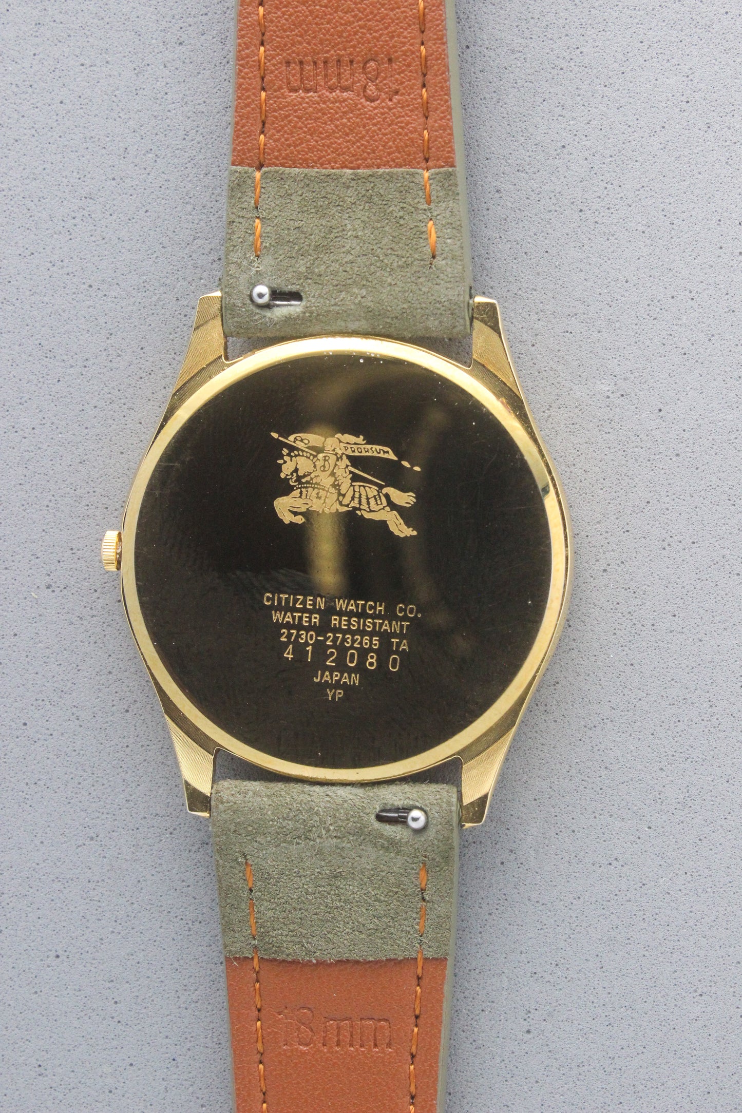 Back view of vintage Burberry wristwatch featuring a polished gold-tone caseback with engraved details, including the iconic Burberrys knight logo and inscriptions. The caseback specifies water resistance and model details: 2280-223465 TA, 412018 0, made in Japan. The watch is paired with a green suede strap and brown leather backing, showcasing aa blend of elegance and durability.