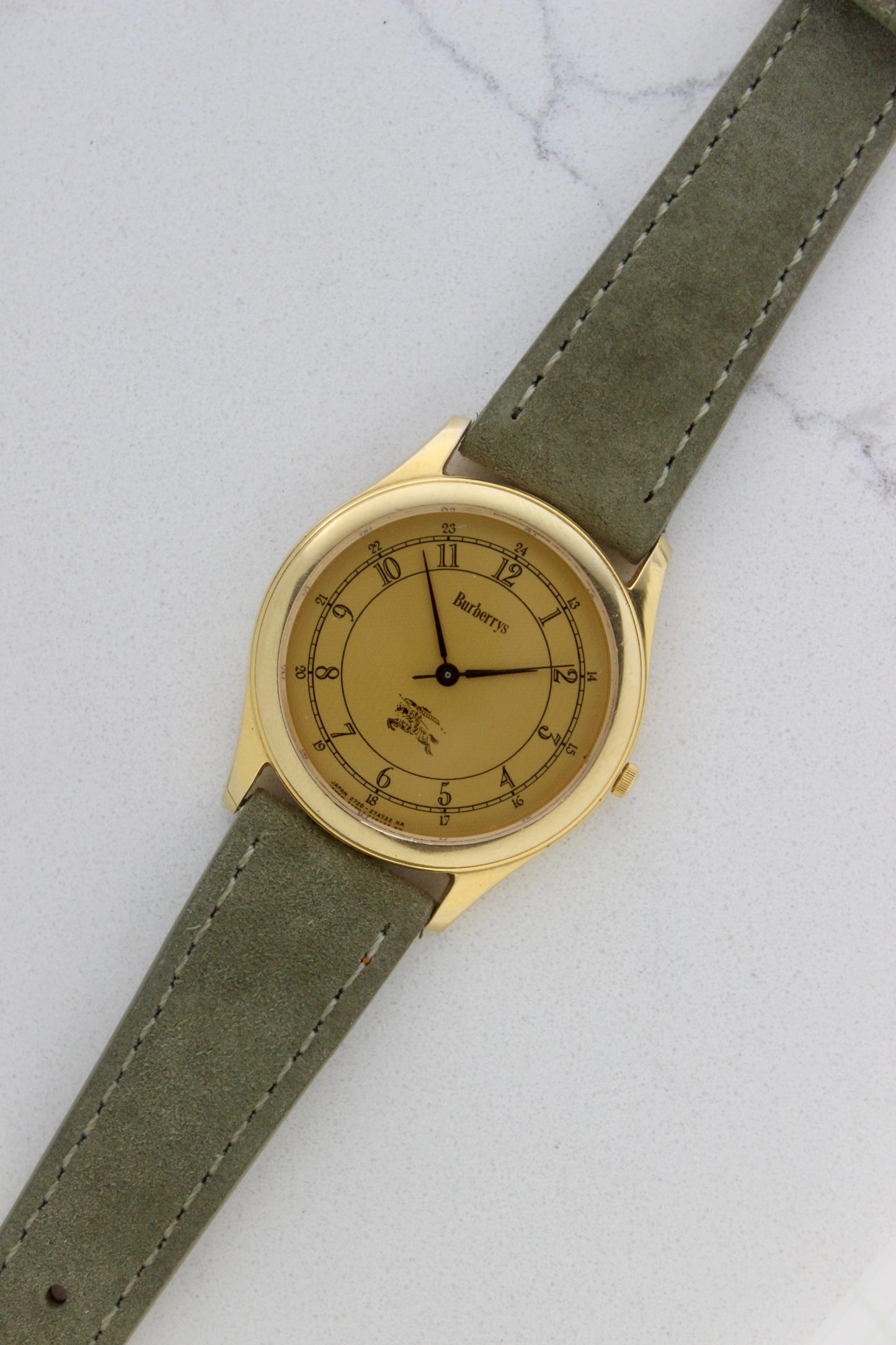 Vintage Burberrys wristwatch with a gold-tone round case, yellow dial featuring black Arabic numerals for both 12-hour and 24-hour markers, and an elegant black logo crest at the 6 o'clock position. The watch is paired with a green suede strap, showcasing a sophisticated and timeless design. Made in Japan, model number 2280-223465 TA, 412018 0.