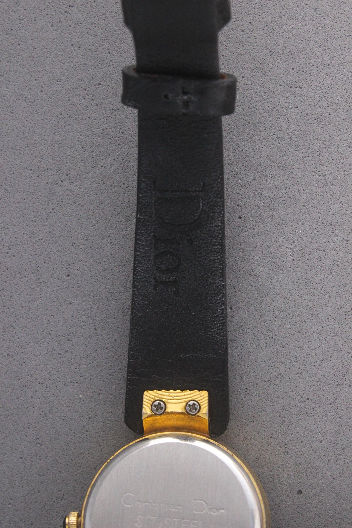 Back view of a Christian Dior Paris vintage watch, ref. 900-11, showcasing a stainless steel case back with engravings and gold-tone accents. Attached to a sleek black leather strap, this timepiece embodies luxury and sophistication, making it a must-have for vintage watch collectors.