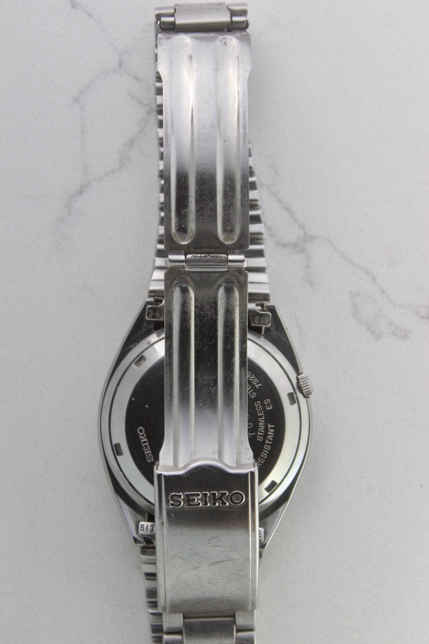The stainless steel bracelet and clasp of a vintage Seiko 5 automatic wristwatch (Reference Number: 7S26-3040). The fold-over clasp is engraved with the Seiko logo and shows visible surface scratches, indicating signs of wear. The original stainless steel bracelet remains intact, with end links marked "B13" and "75-E". This Japan-exclusive mid-1980s model is in good condition with expected wear for its age.