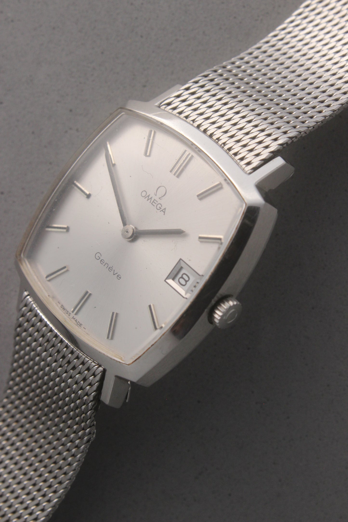 Vintage Omega Genève men’s wristwatch, reference number 132.0052, featuring a sleek silver square case, minimalist silver-tone dial, date display at 3 o’clock, and an elegant stainless steel mesh bracelet. A timeless Swiss-made classic, perfect for collectors and enthusiasts of luxury timepieces.