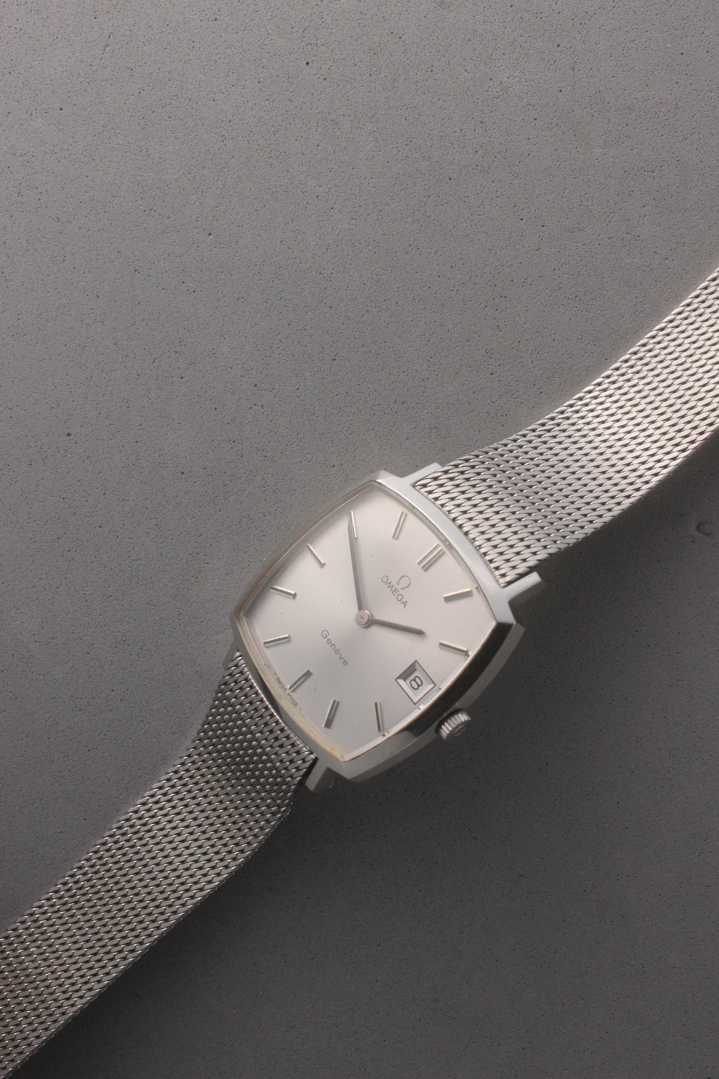 Vintage Omega Genève men’s wristwatch, reference number 132.0052, featuring a sleek silver square case, minimalist silver-tone dial, date display at 3 o’clock, and an elegant stainless steel mesh bracelet. A timeless Swiss-made classic, perfect for collectors and enthusiasts of luxury timepieces.