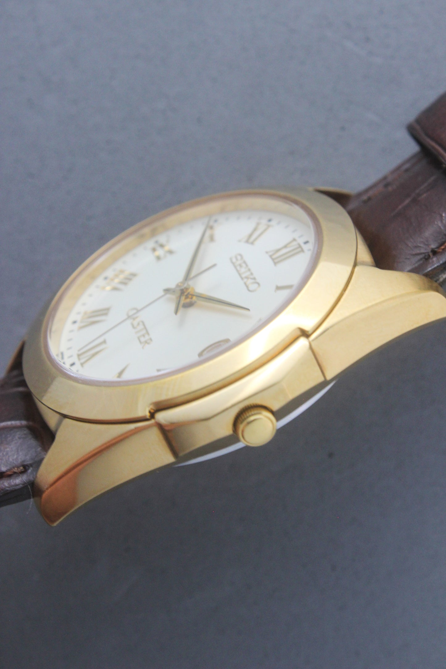 A detailed front view of the Seiko Caster
8F32-0140 watch featuring a gold-tone case, Roman numeral markers, and a date window at 3 o'clock. The cream-colored dial is complemented by classic black hands, encapsulating an elegant vintage aesthetic.
Paired with a genuine brown leather strap.