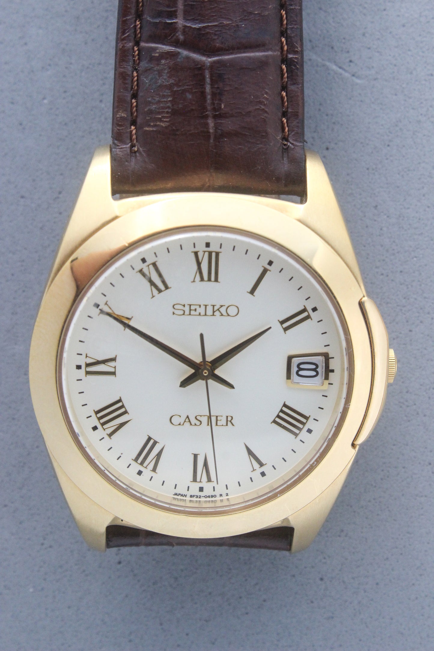 A detailed front view of the Seiko Caster
8F32-0140 watch featuring a gold-tone case, Roman numeral markers, and a date window at 3 o'clock. The cream-colored dial is complemented by classic black hands, encapsulating an elegant vintage aesthetic.
Paired with a genuine brown leather strap.