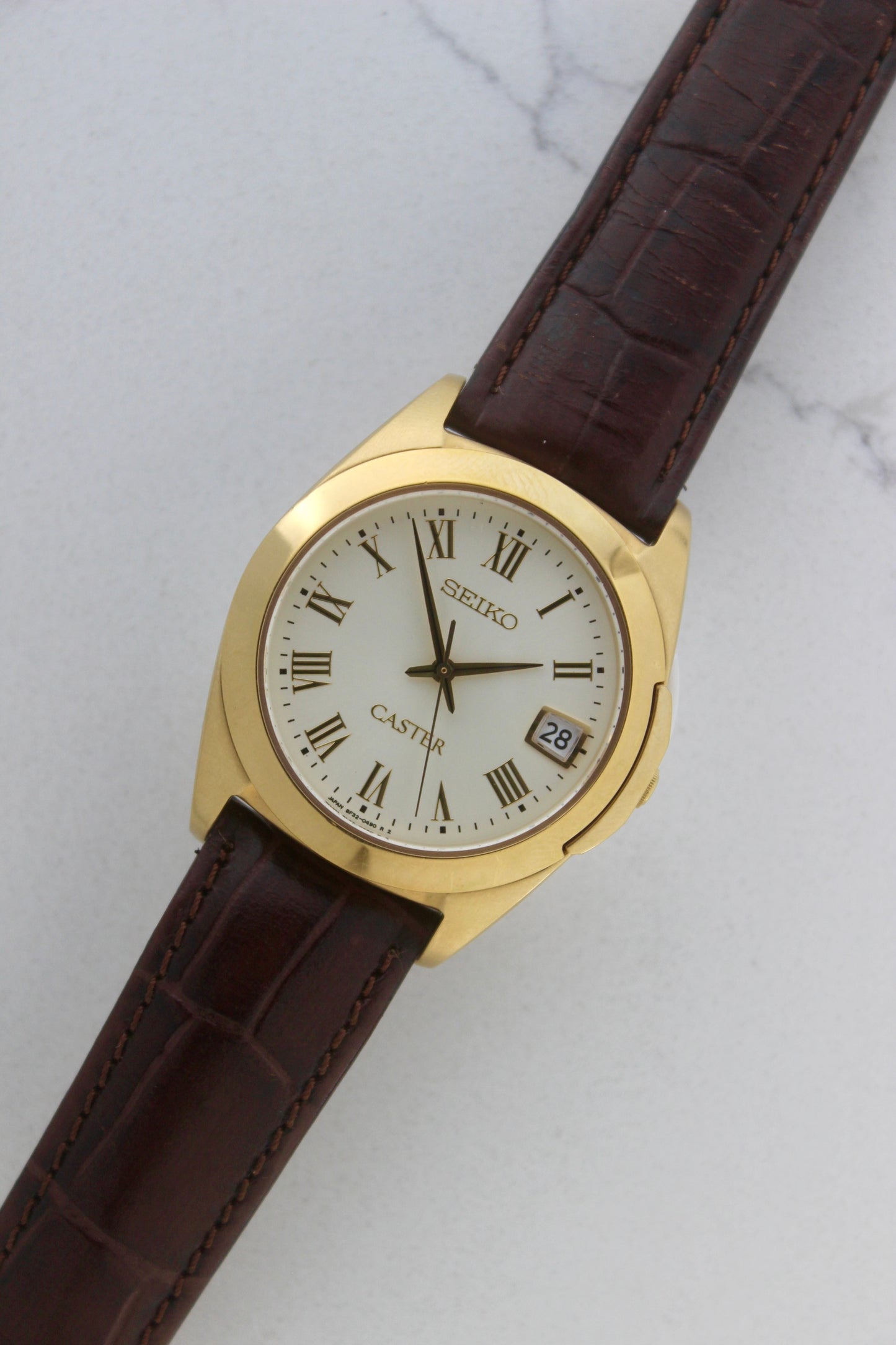 A detailed front view of the Seiko Caster
8F32-0140 watch featuring a gold-tone case, Roman numeral markers, and a date window at 3 o'clock. The cream-colored dial is complemented by classic black hands, encapsulating an elegant vintage aesthetic.
Paired with a genuine brown leather strap.