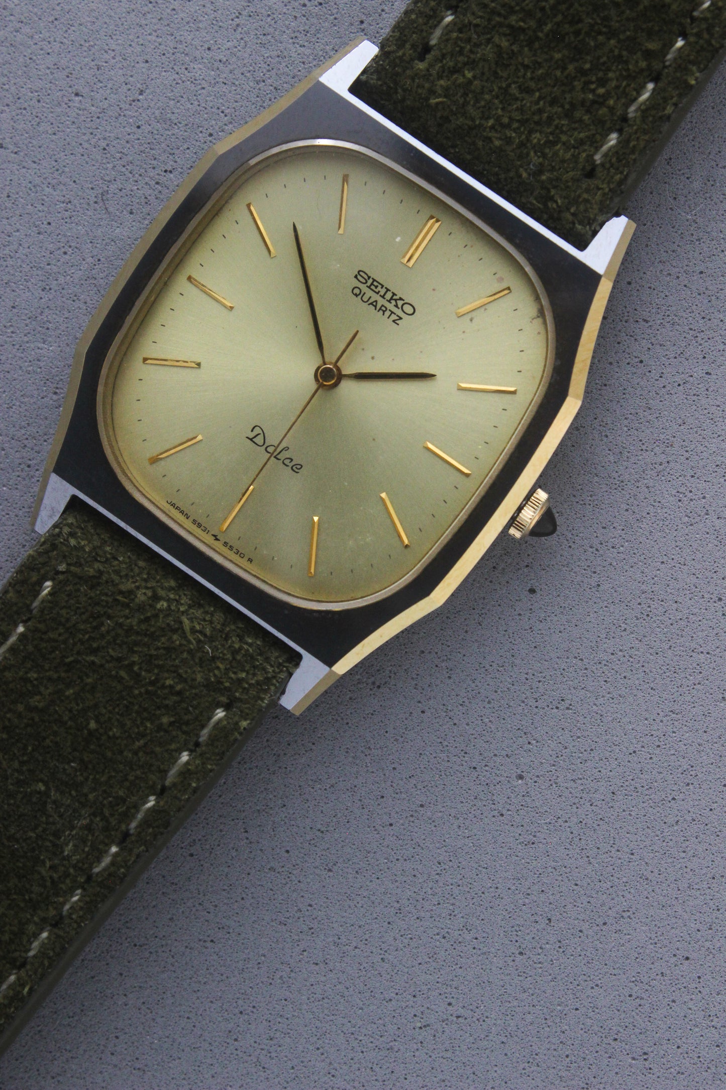A close-up of the Seiko Dolce 5931-5420
watch featuring a rectangular stainless steel case with a gold-plated bezel, a champagne dial, and gold hour indices. Paired with an elegant green suede strap, this minimalist design showcases timeless sophistication.
