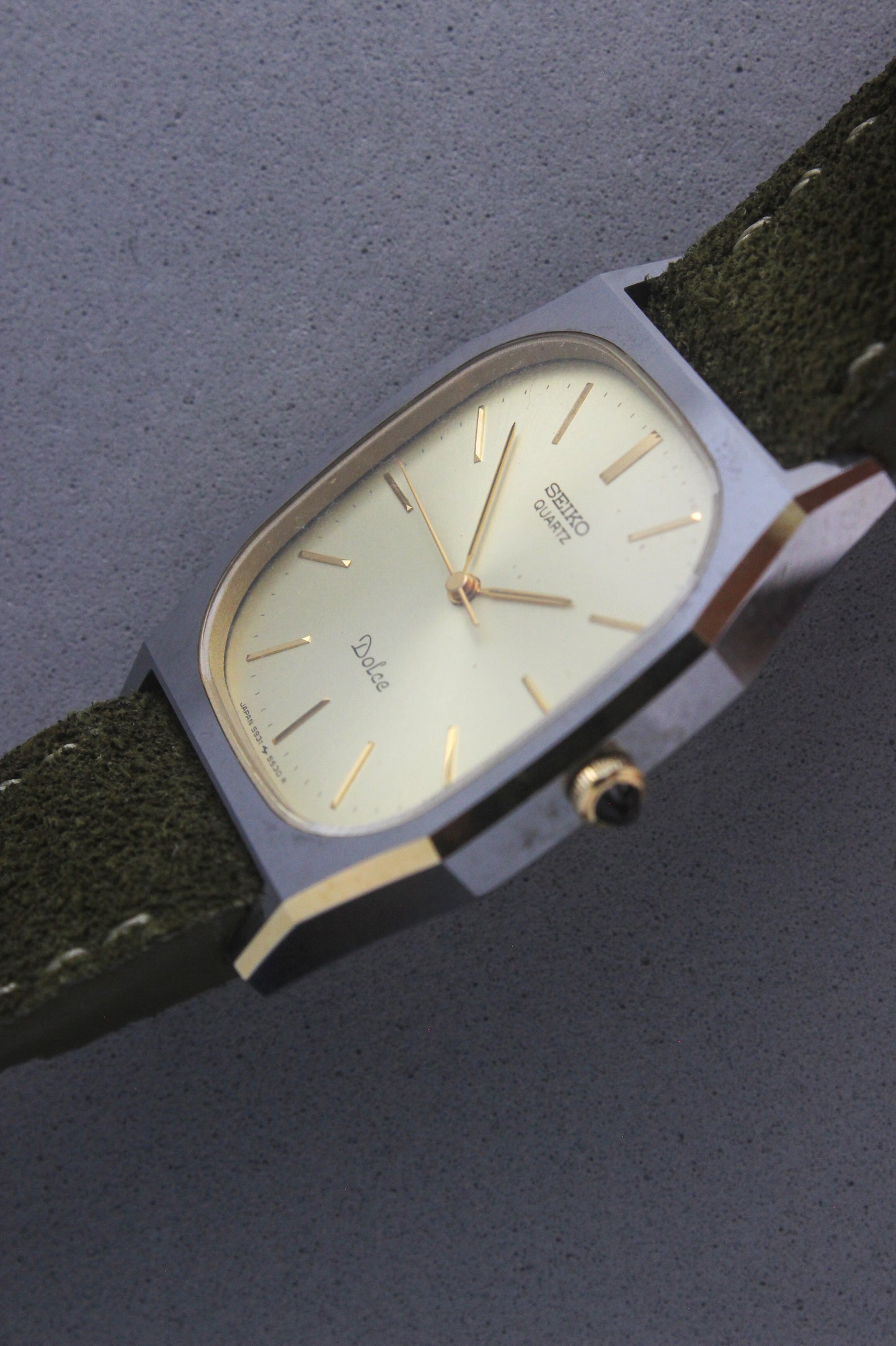 A close-up of the Seiko Dolce 5931-5420
watch featuring a rectangular stainless steel case with a gold-plated bezel, a champagne dial, and gold hour indices. Paired with an elegant green suede strap, this minimalist design showcases timeless sophistication.