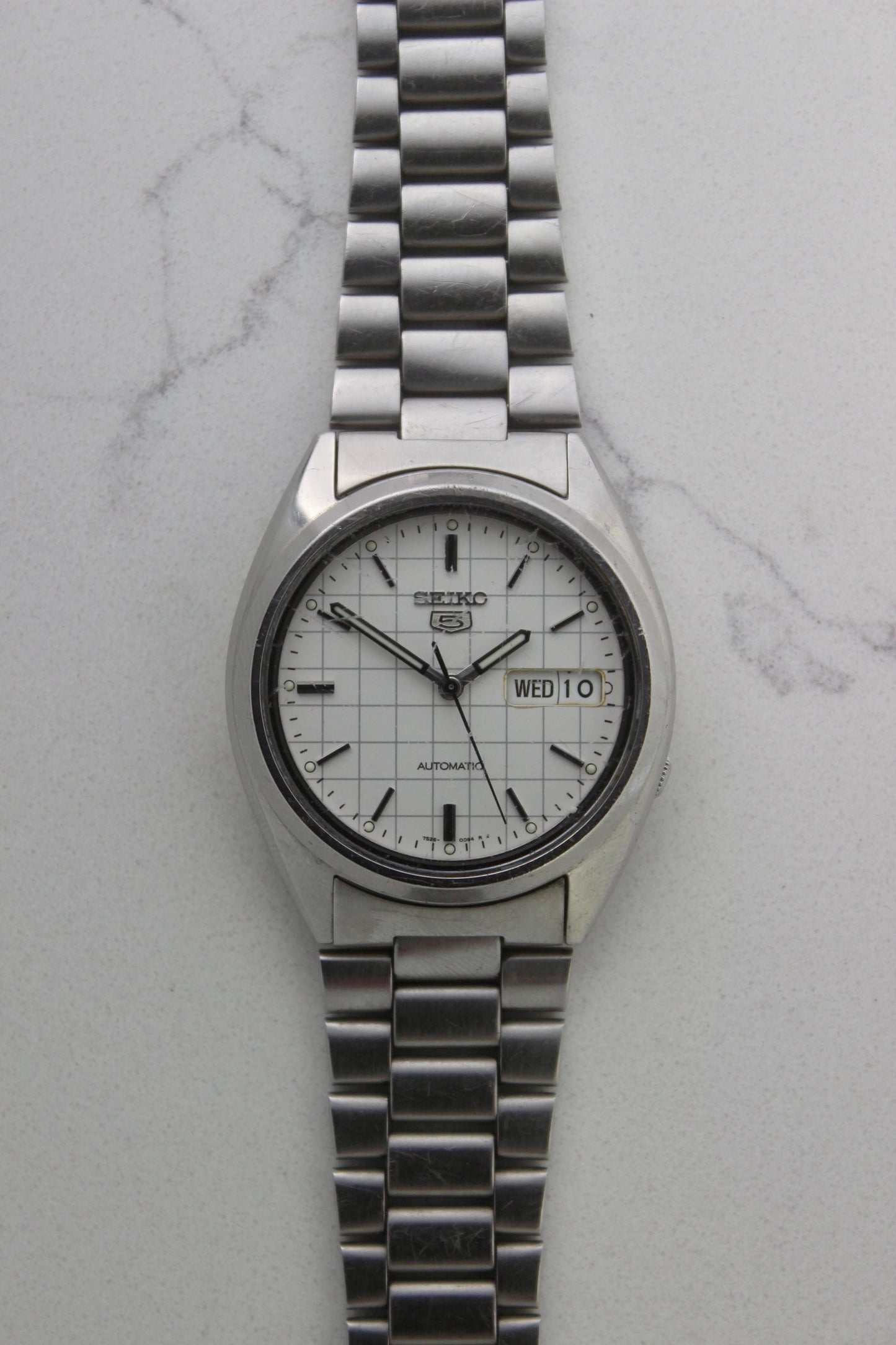 A vintage Seiko 5 automatic wristwatch (Reference Number: 7S26-3040) with a stainless steel case and bracelet. The watch features a 36mm white dial with a grid-painted design and a day-date function at the 3 o’clock position. The stainless steel bezel is fixed, complementing the original bracelet, which fits up to a 7-inch wrist. This Japan-exclusive mid-1980s model is in good condition, showing signs of wear on the bracelet, buckle, and crystal.