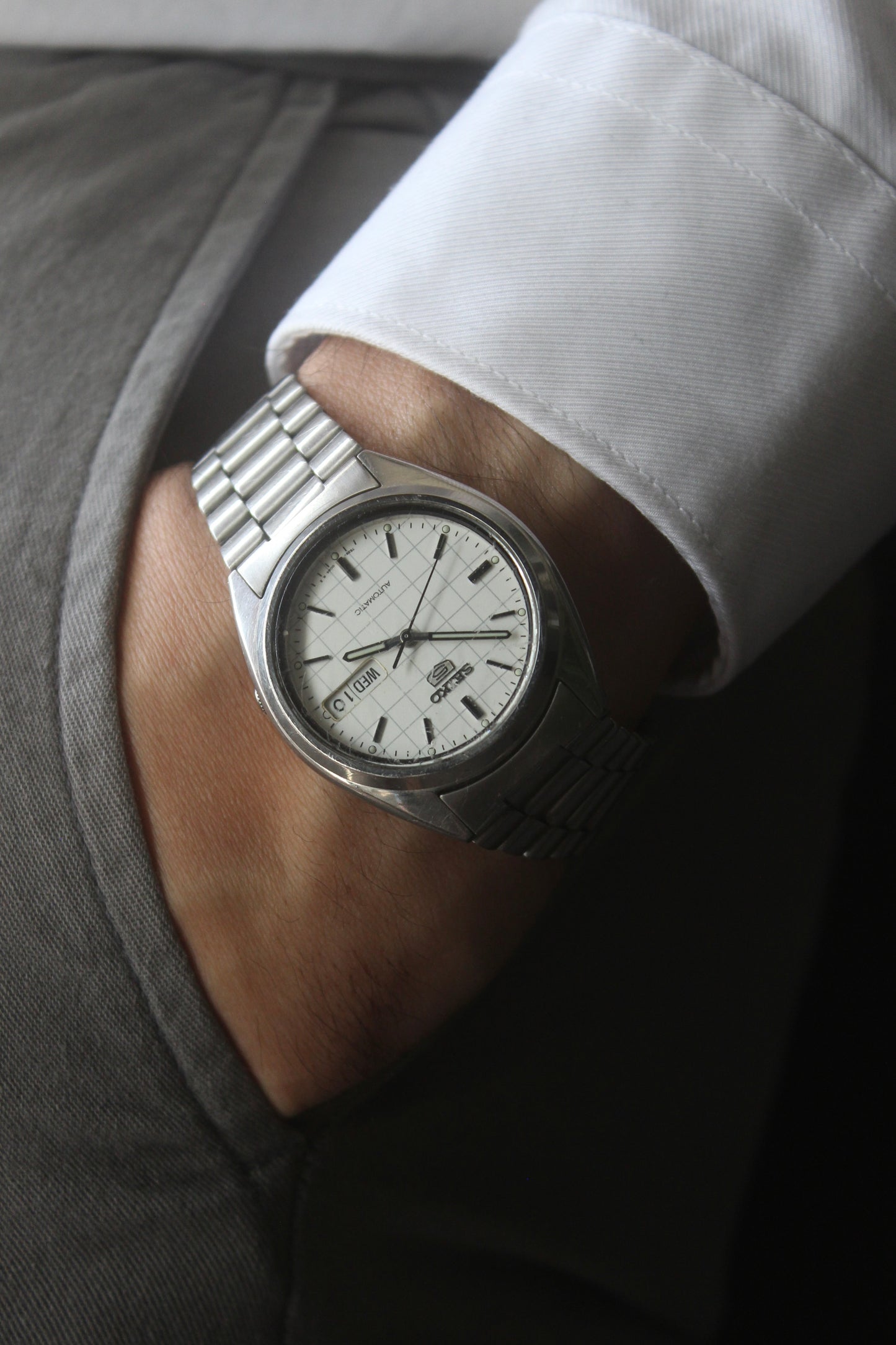 A vintage Seiko 5 automatic wristwatch (Reference Number: 7S26-3040) with a stainless steel case and bracelet. The watch features a 36mm white dial with a grid-painted design and a day-date function at the 3 o’clock position. The stainless steel bezel is fixed, complementing the original bracelet, which fits up to a 7-inch wrist. This Japan-exclusive mid-1980s model is in good condition, showing signs of wear on the bracelet, buckle, and crystal.