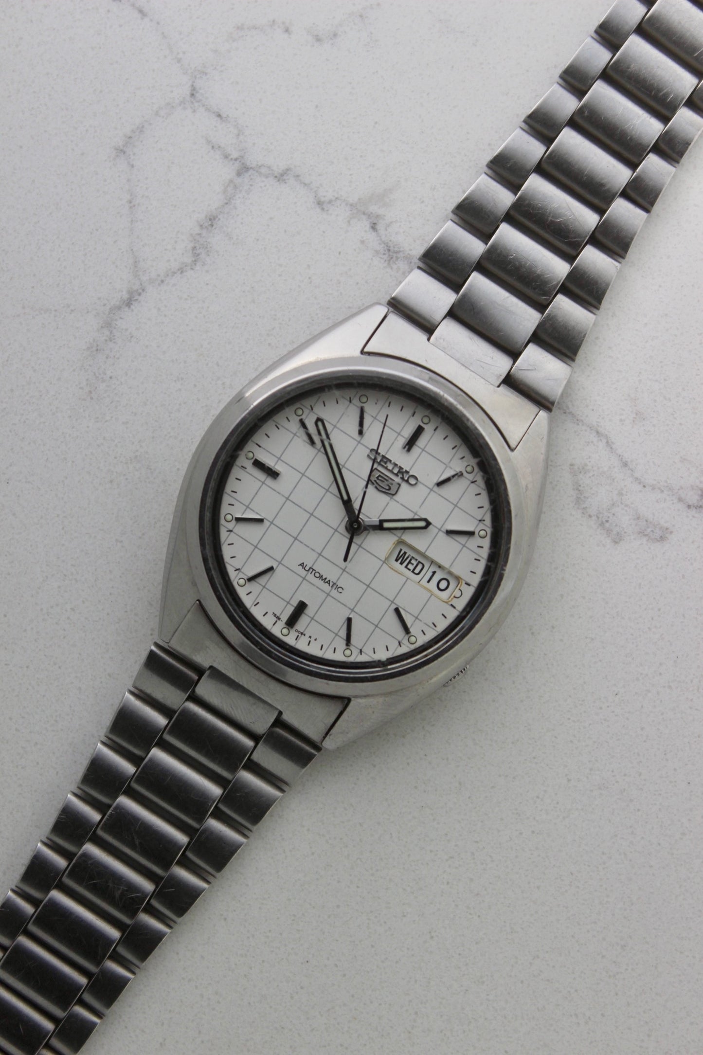 A vintage Seiko 5 automatic wristwatch (Reference Number: 7S26-3040) with a stainless steel case and bracelet. The watch features a 36mm white dial with a grid-painted design and a day-date function at the 3 o’clock position. The stainless steel bezel is fixed, complementing the original bracelet, which fits up to a 7-inch wrist. This Japan-exclusive mid-1980s model is in good condition, showing signs of wear on the bracelet, buckle, and crystal.