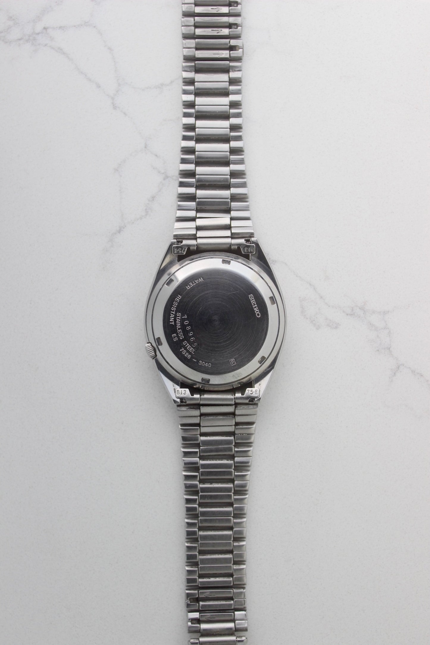 The case back of a vintage Seiko 5 automatic wristwatch (Reference Number: 7S26-3040) made of stainless steel. The screw-down case back is engraved with Seiko branding, water resistance details, and serial numbers, showing visible signs of wear including surface scratches. The original stainless steel bracelet is attached, featuring end links marked "B13" and "75-E". This Japan-exclusive mid-1980s model is in good condition with expected wear for its age.