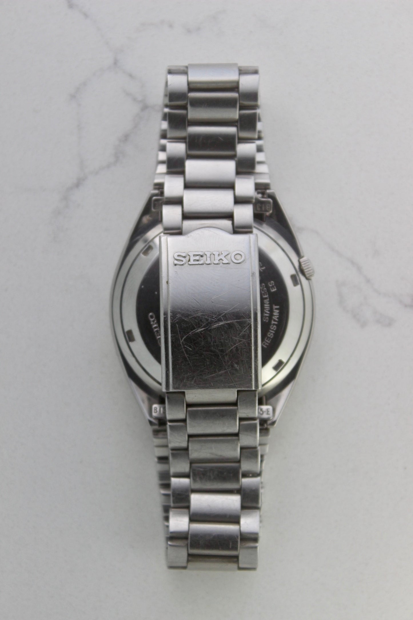 The stainless steel bracelet and clasp of a vintage Seiko 5 automatic wristwatch (Reference Number: 7S26-3040). The fold-over clasp is engraved with the Seiko logo and shows visible surface scratches, indicating signs of wear. The original stainless steel bracelet remains intact, with end links marked "B13" and "75-E". This Japan-exclusive mid-1980s model is in good condition with expected wear for its age.