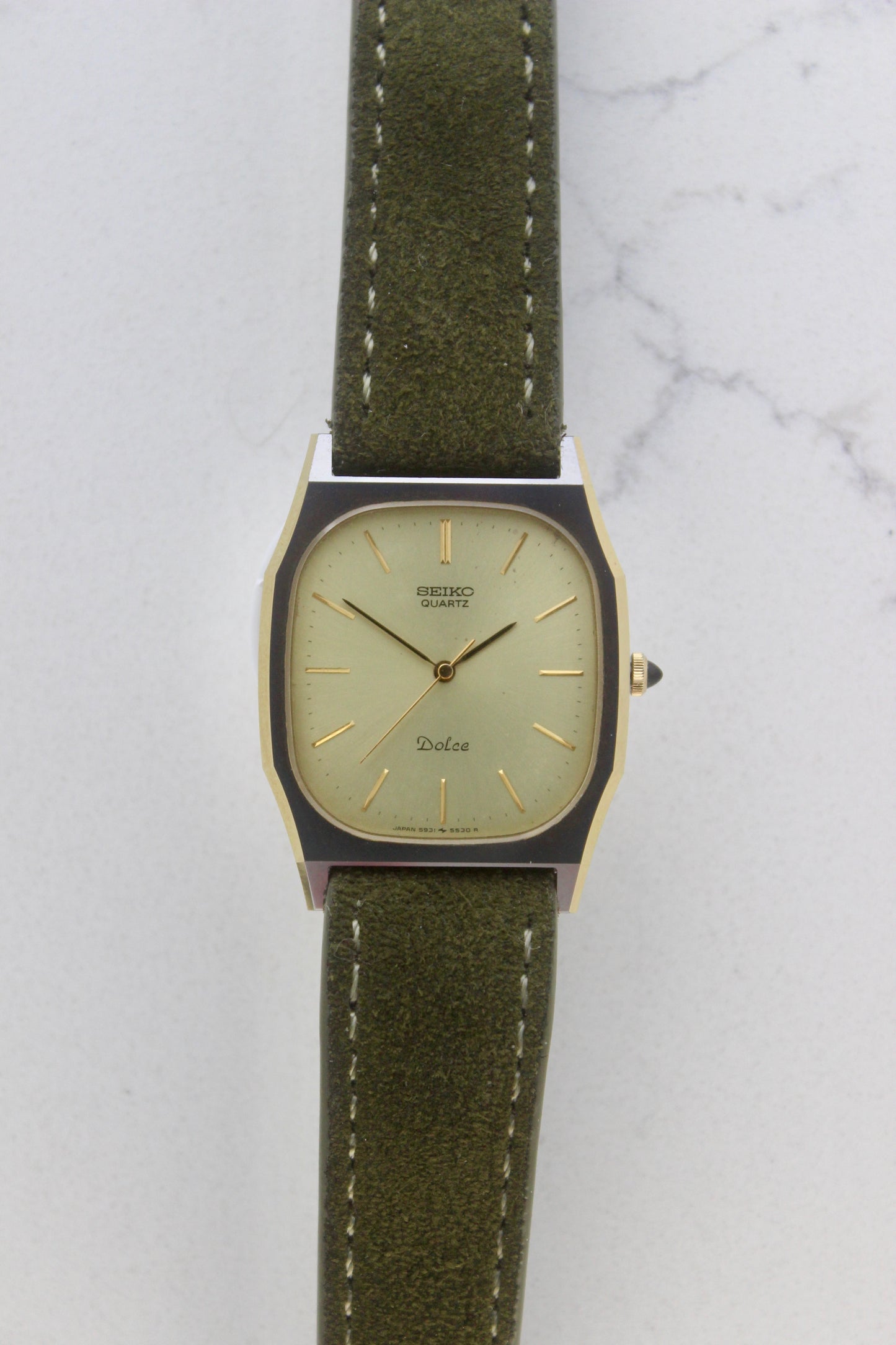 A close-up of the Seiko Dolce 5931-5420
watch featuring a rectangular stainless steel case with a gold-plated bezel, a champagne dial, and gold hour indices. Paired with an elegant green suede strap, this minimalist design showcases timeless sophistication.