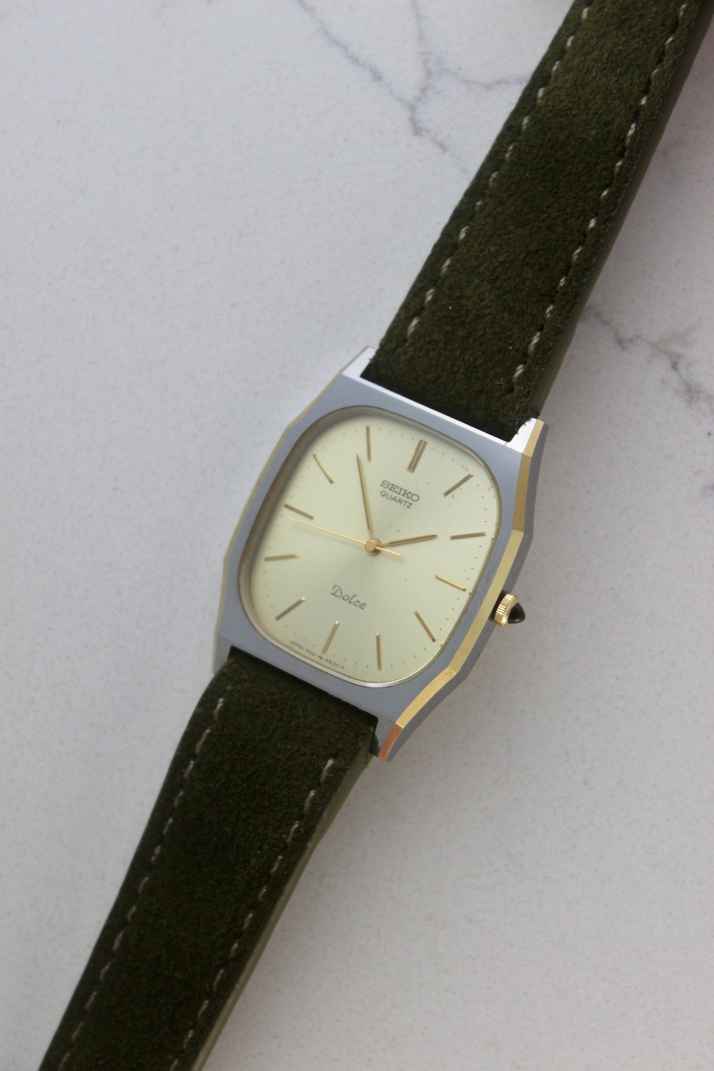 A close-up of the Seiko Dolce 5931-5420
watch featuring a rectangular stainless steel case with a gold-plated bezel, a champagne dial, and gold hour indices. Paired with an elegant green suede strap, this minimalist design showcases timeless sophistication.