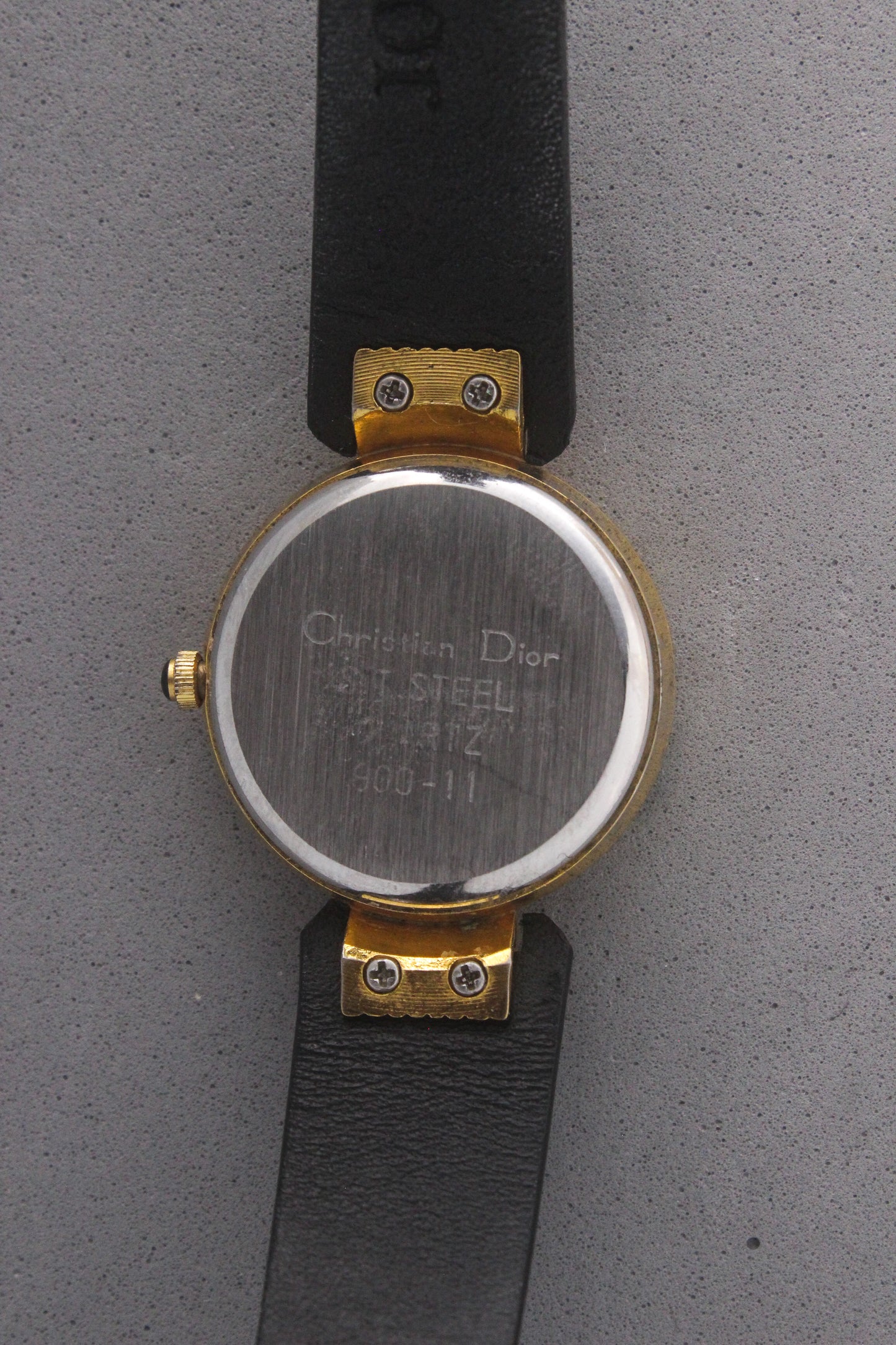 Back view of a Christian Dior Paris vintage watch, ref. 900-11, showcasing a stainless steel case back with engravings and gold-tone accents. Attached to a sleek black leather strap, this timepiece embodies luxury and sophistication, making it a must-have for vintage watch collectors.