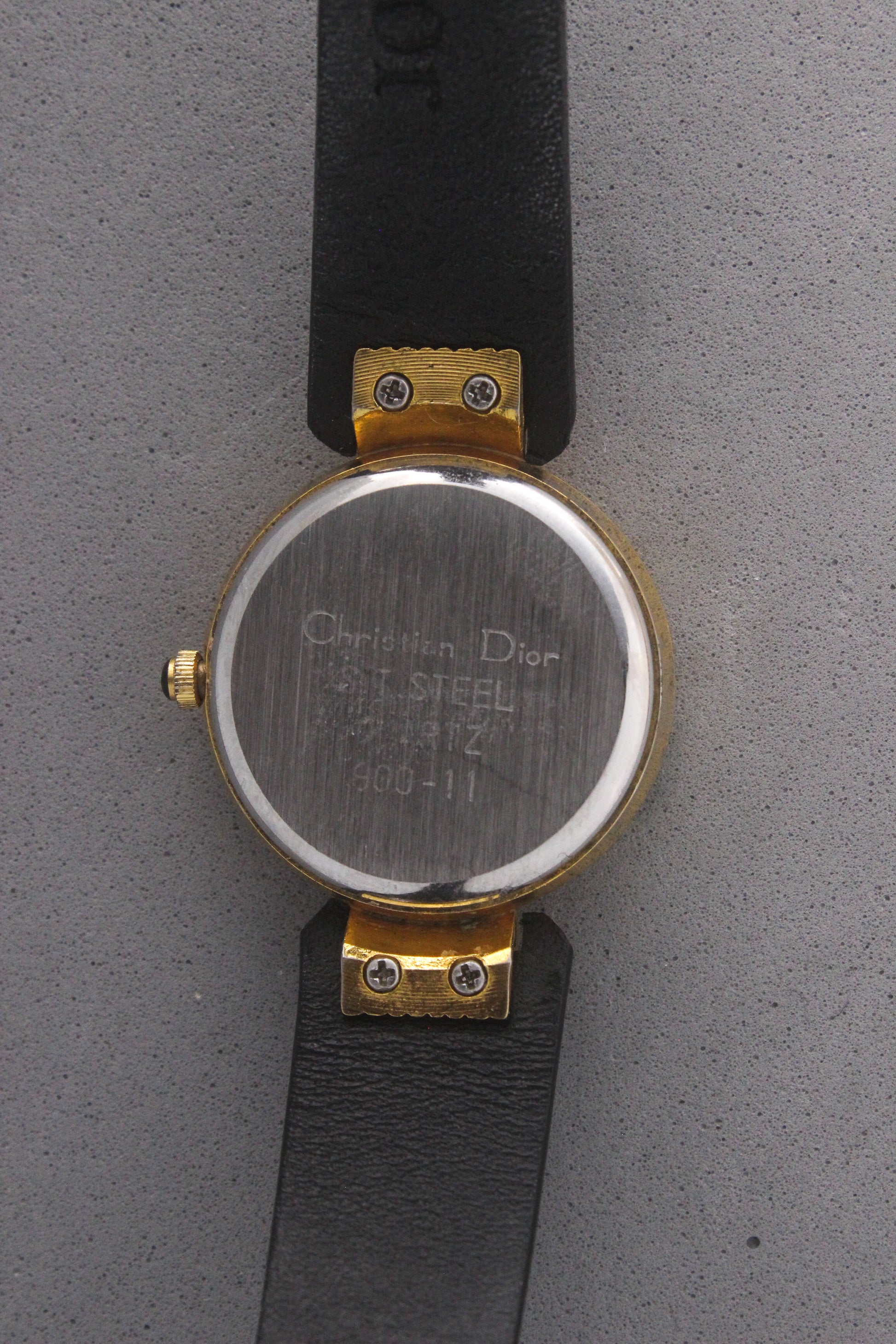 Back view of a Christian Dior Paris vintage watch, ref. 900-11, showcasing a stainless steel case back with engravings and gold-tone accents. Attached to a sleek black leather strap, this timepiece embodies luxury and sophistication, making it a must-have for vintage watch collectors.