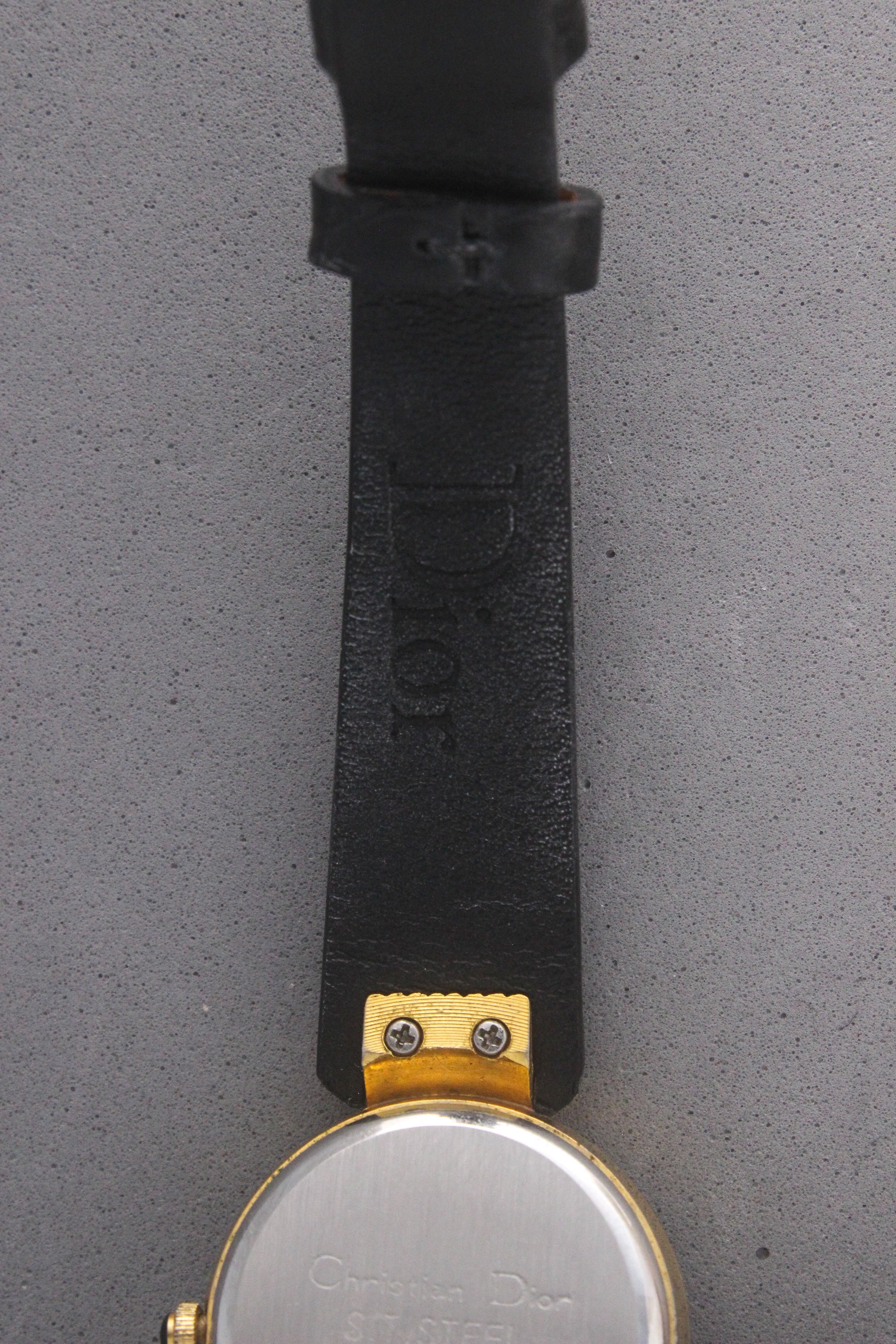 Back view of a Christian Dior Paris vintage watch, ref. 900-11, showcasing a stainless steel case back with engravings and gold-tone accents. Attached to a sleek black leather strap, this timepiece embodies luxury and sophistication, making it a must-have for vintage watch collectors.