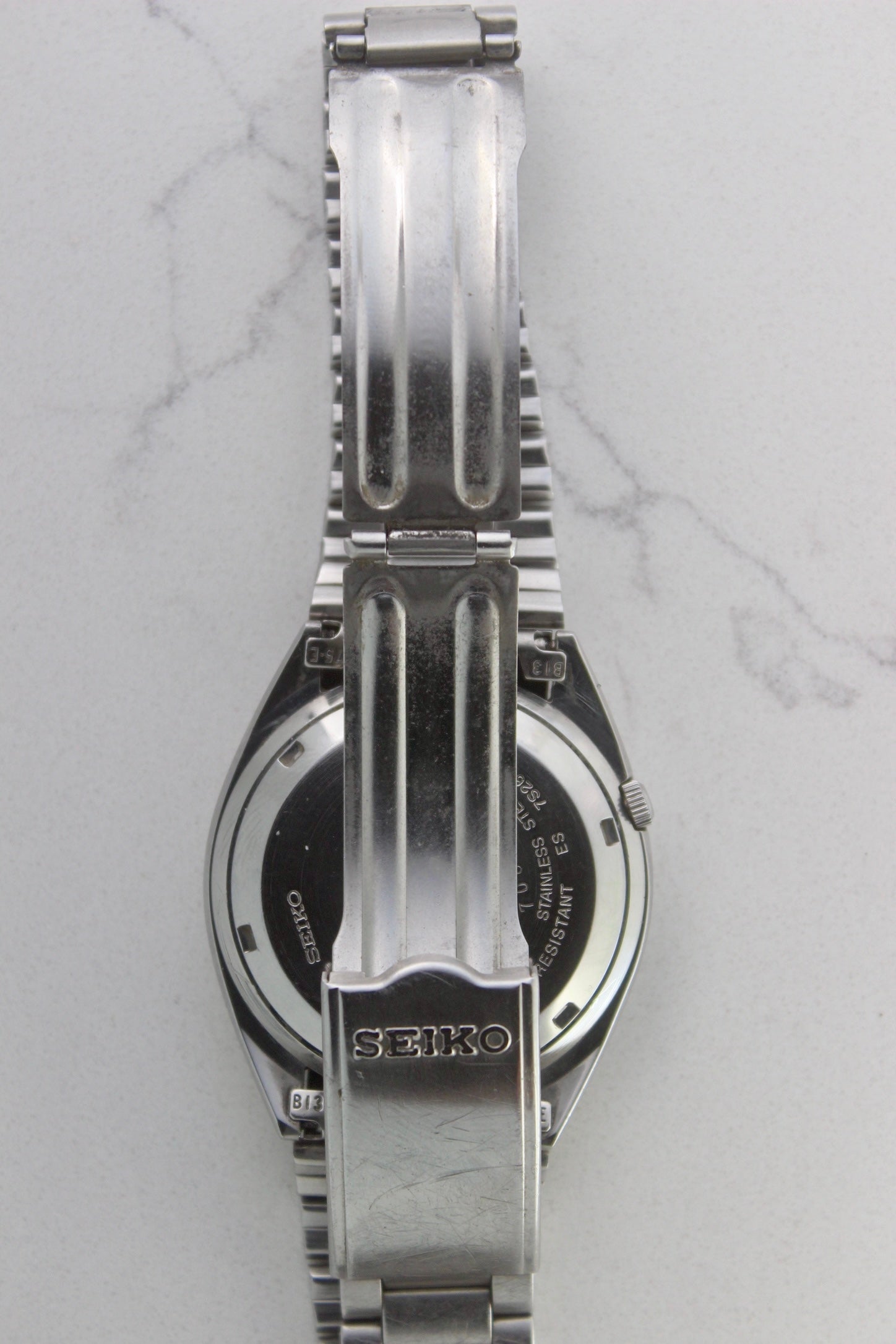 The stainless steel bracelet and clasp of a vintage Seiko 5 automatic wristwatch (Reference Number: 7S26-3040). The fold-over clasp is engraved with the Seiko logo and shows visible surface scratches, indicating signs of wear. The original stainless steel bracelet remains intact, with end links marked "B13" and "75-E". This Japan-exclusive mid-1980s model is in good condition with expected wear for its age.