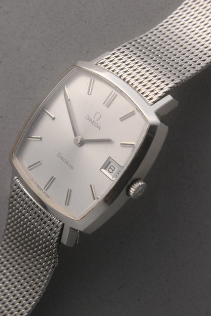 Vintage Omega Genève men’s wristwatch, reference number 132.0052, featuring a sleek silver square case, minimalist silver-tone dial, date display at 3 o’clock, and an elegant stainless steel mesh bracelet. A timeless Swiss-made classic, perfect for collectors and enthusiasts of luxury timepieces.