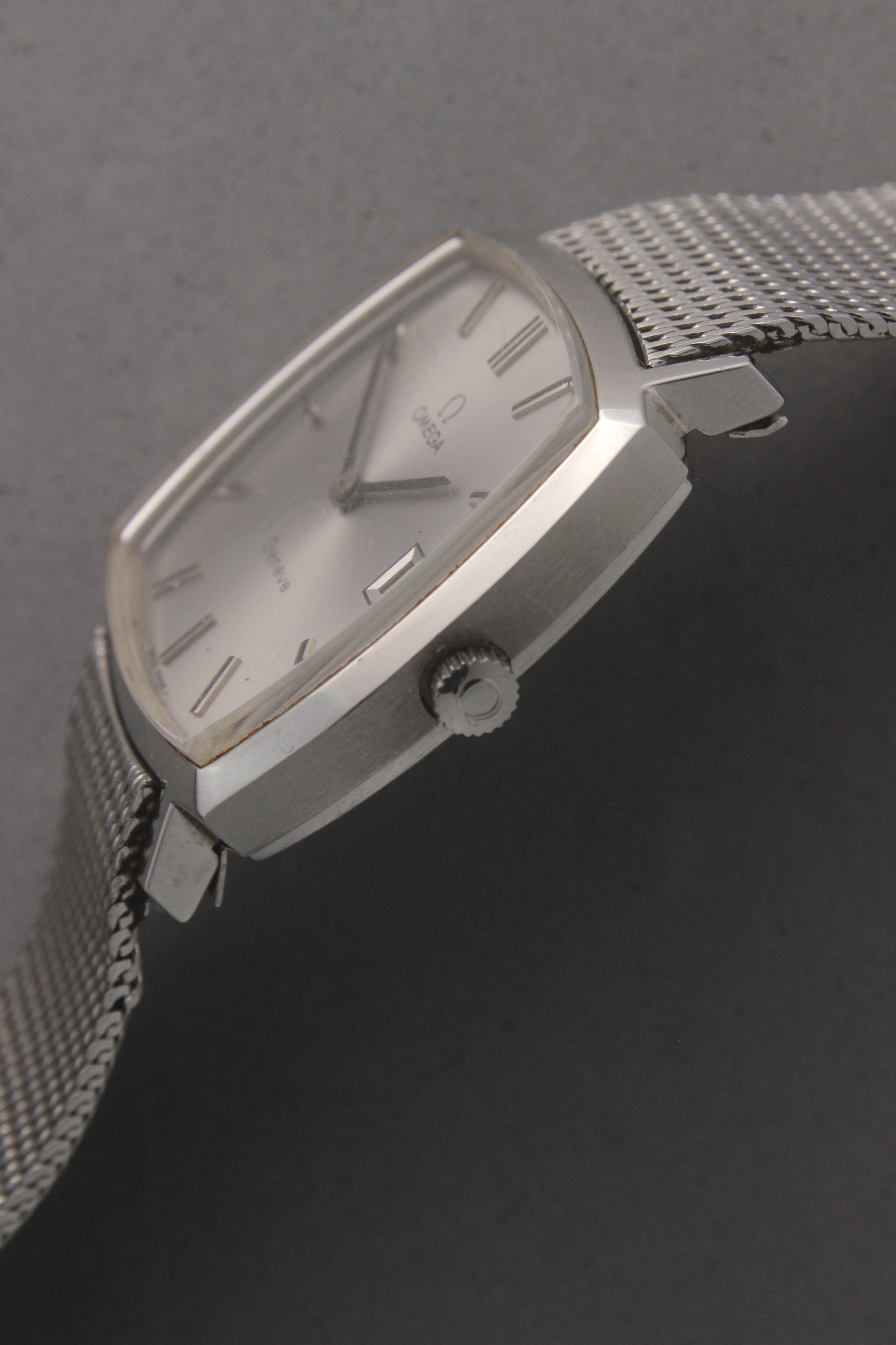 Vintage Omega Genève men’s wristwatch, reference number 132.0052, featuring a sleek silver square case, minimalist silver-tone dial, date display at 3 o’clock, and an elegant stainless steel mesh bracelet. A timeless Swiss-made classic, perfect for collectors and enthusiasts of luxury timepieces.
