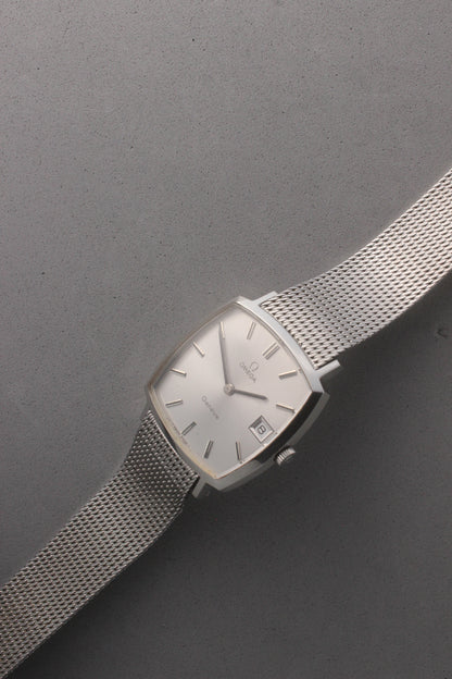 Vintage Omega Genève men’s wristwatch, reference number 132.0052, featuring a sleek silver square case, minimalist silver-tone dial, date display at 3 o’clock, and an elegant stainless steel mesh bracelet. A timeless Swiss-made classic, perfect for collectors and enthusiasts of luxury timepieces.