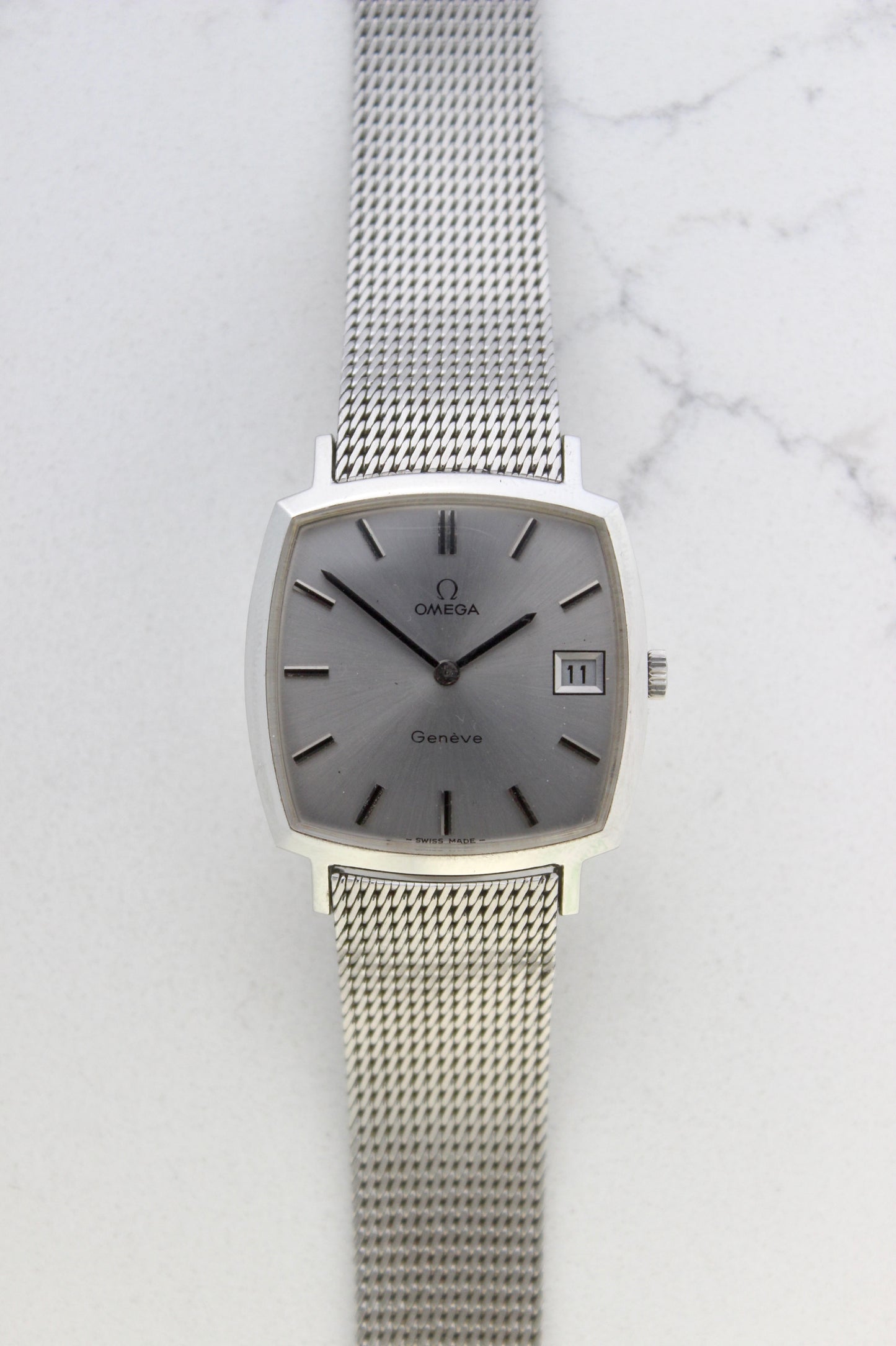Vintage Omega Genève men’s wristwatch, reference number 132.0052, featuring a sleek silver square case, minimalist silver-tone dial, date display at 3 o’clock, and an elegant stainless steel mesh bracelet. A timeless Swiss-made classic, perfect for collectors and enthusiasts of luxury timepieces.