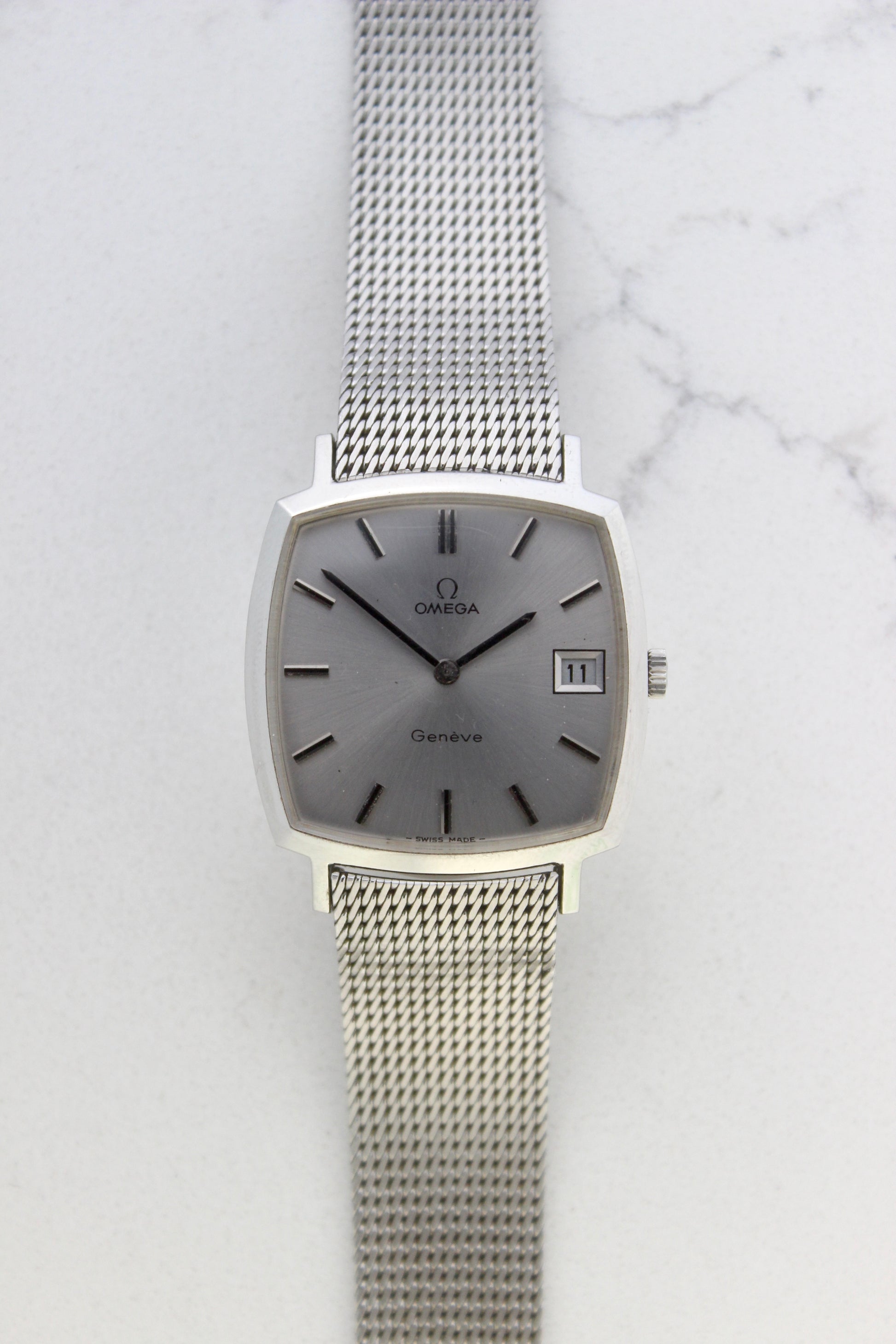 Vintage Omega Genève men’s wristwatch, reference number 132.0052, featuring a sleek silver square case, minimalist silver-tone dial, date display at 3 o’clock, and an elegant stainless steel mesh bracelet. A timeless Swiss-made classic, perfect for collectors and enthusiasts of luxury timepieces.