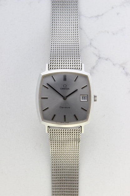 Vintage Omega Genève men’s wristwatch, reference number 132.0052, featuring a sleek silver square case, minimalist silver-tone dial, date display at 3 o’clock, and an elegant stainless steel mesh bracelet. A timeless Swiss-made classic, perfect for collectors and enthusiasts of luxury timepieces.