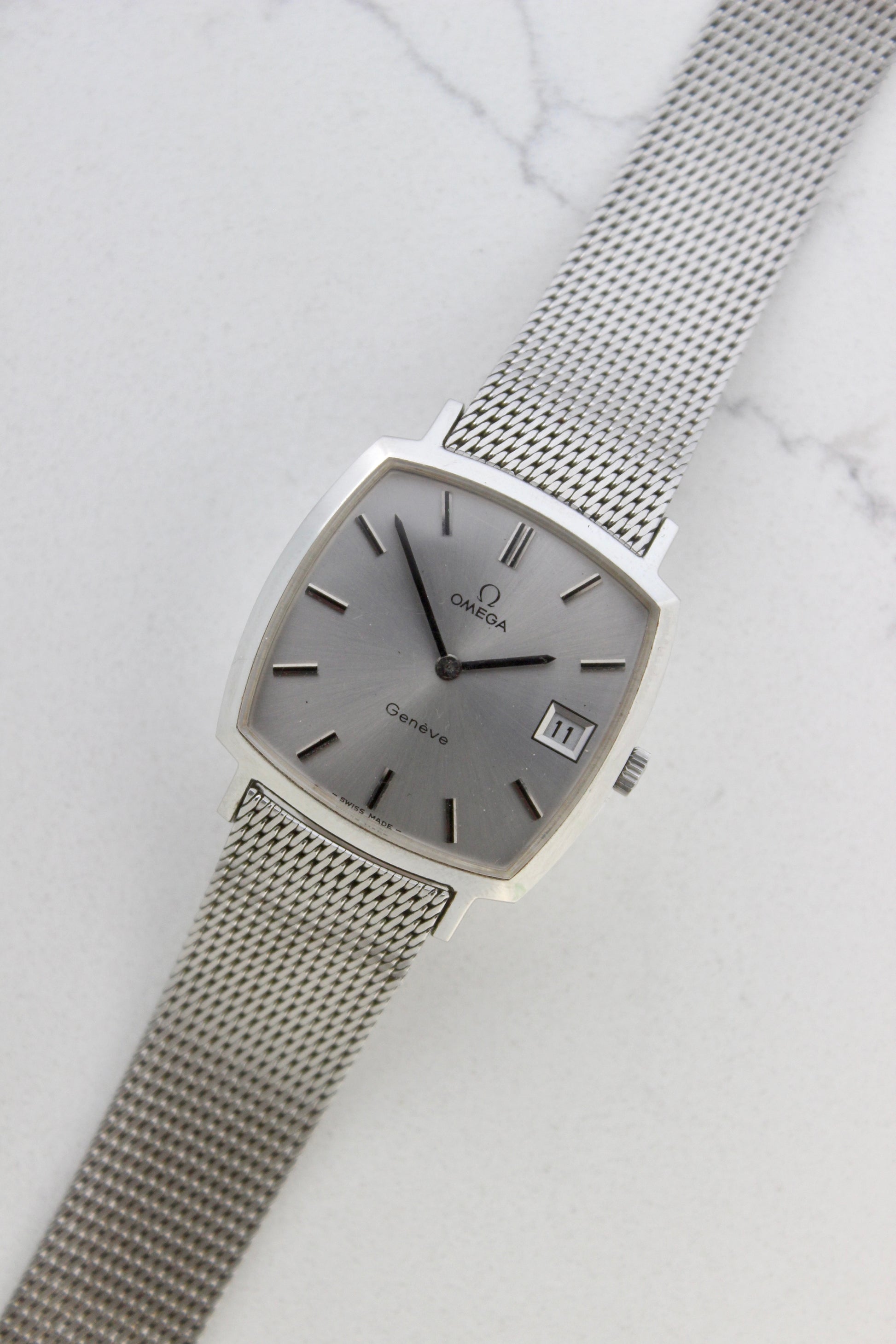 Vintage Omega Genève men’s wristwatch, reference number 132.0052, featuring a sleek silver square case, minimalist silver-tone dial, date display at 3 o’clock, and an elegant stainless steel mesh bracelet. A timeless Swiss-made classic, perfect for collectors and enthusiasts of luxury timepieces.