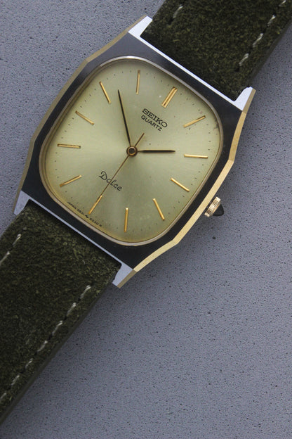 A close-up of the Seiko Dolce 5931-5420
watch featuring a rectangular stainless steel case with a gold-plated bezel, a champagne dial, and gold hour indices. Paired with an elegant green suede strap, this minimalist design showcases timeless sophistication.