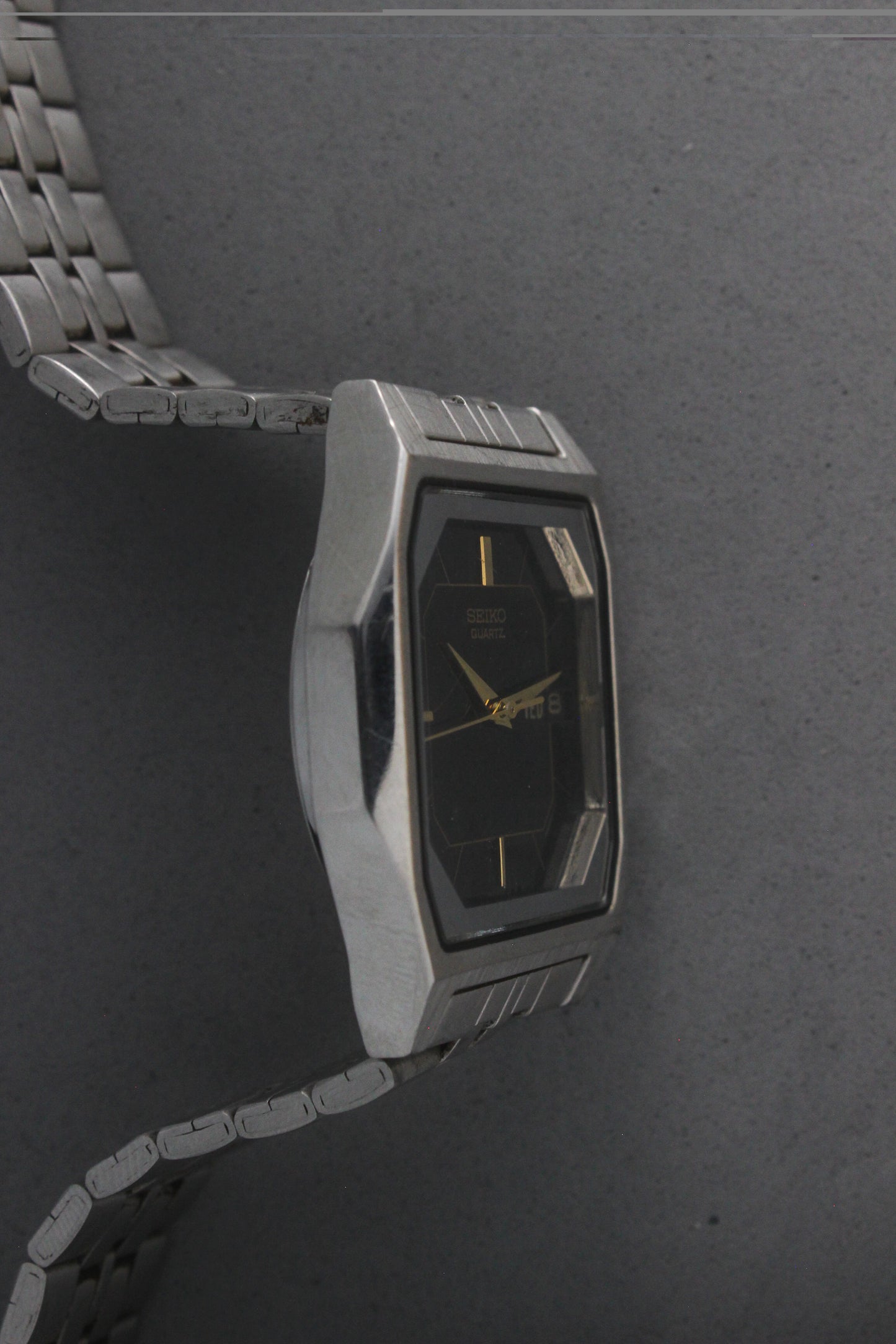 A close-up of the Seiko Quartz 7N43-5011, featuring a rectangular stainless steel case, black dial with gold accents, and a day-date window at 3 o'clock. The watch is paired with a stainless steel bracelet, exuding a timeless and versatile design.
