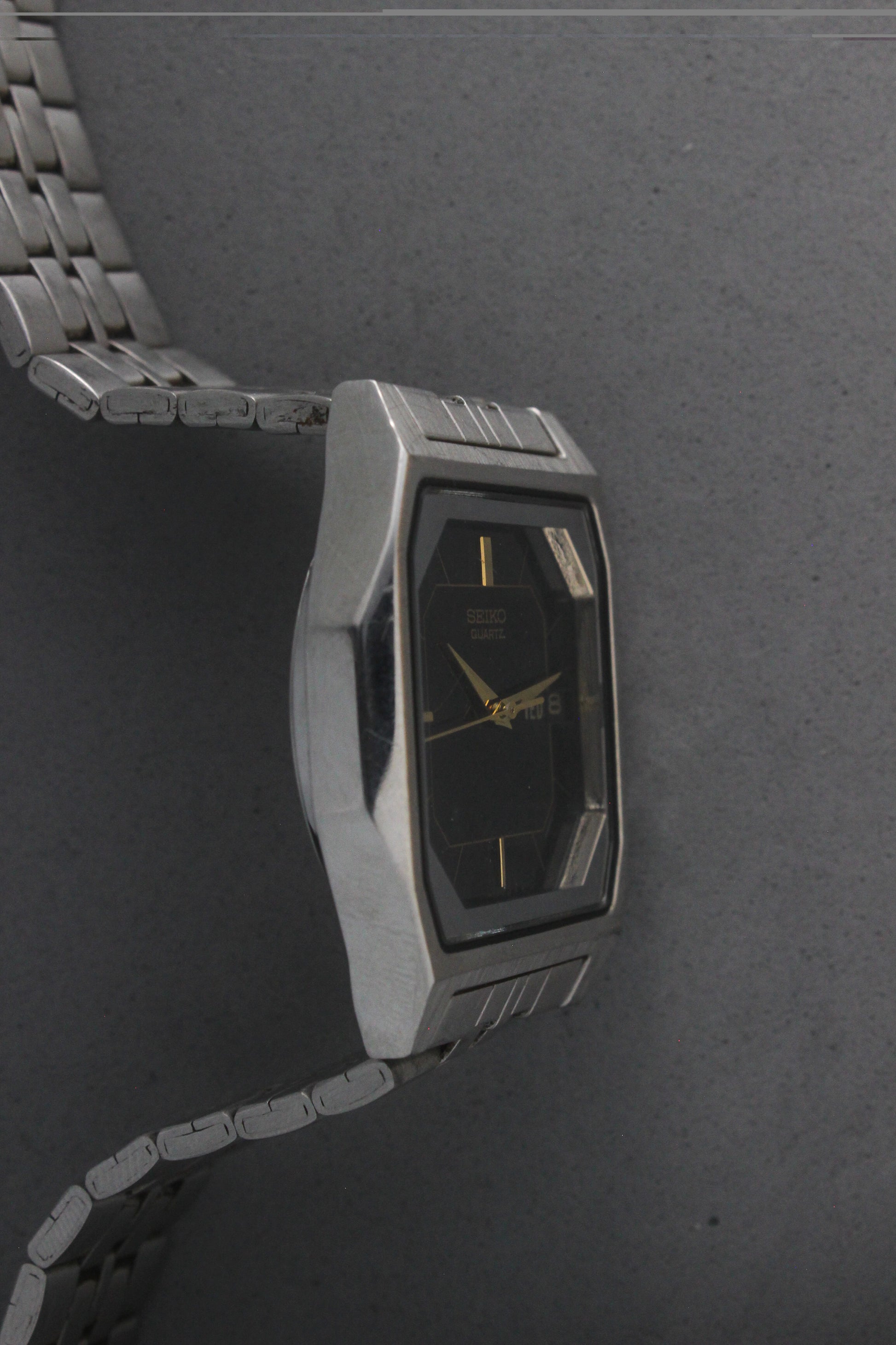 A close-up of the Seiko Quartz 7N43-5011, featuring a rectangular stainless steel case, black dial with gold accents, and a day-date window at 3 o'clock. The watch is paired with a stainless steel bracelet, exuding a timeless and versatile design.
