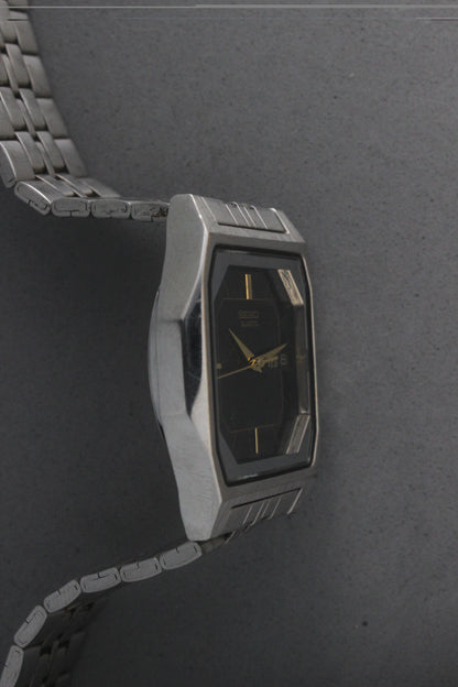 A close-up of the Seiko Quartz 7N43-5011, featuring a rectangular stainless steel case, black dial with gold accents, and a day-date window at 3 o'clock. The watch is paired with a stainless steel bracelet, exuding a timeless and versatile design.