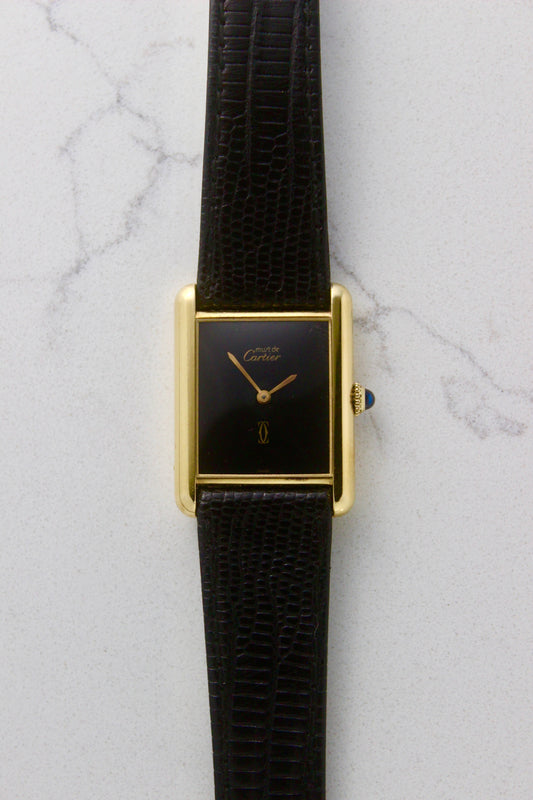 A Must de Cartier Tank watch with a yellow gold-plated sterling silver case, deep black ghost dial, and gold-tone hands. The watch is paired with a black lizard strap and features a cabochon crown. Photographed on a marble surface.