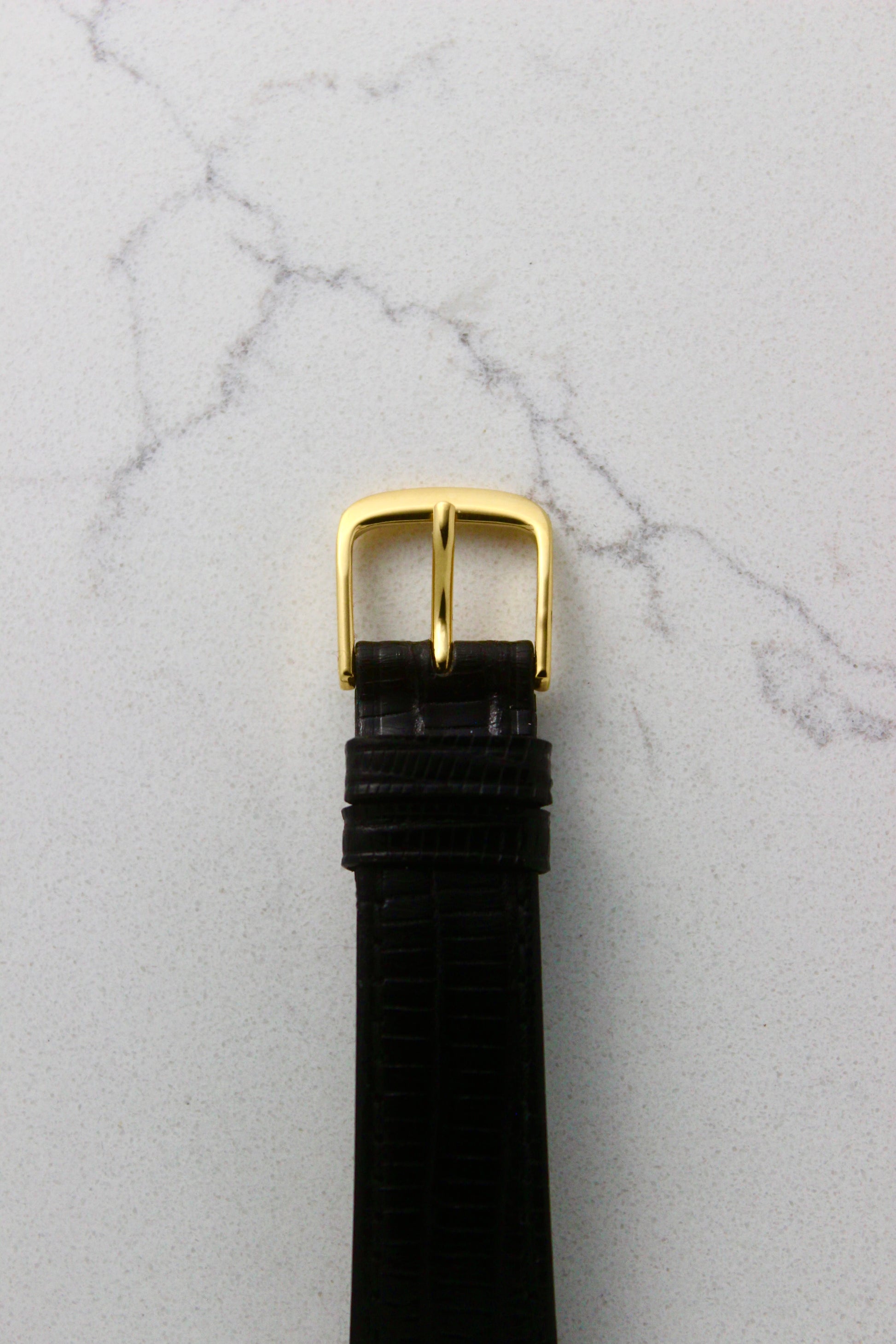 Close-up of a black lizard leather watch strap with a gold-tone buckle, photographed on a marble surface. The strap features two matching leather keepers for a secure fit.