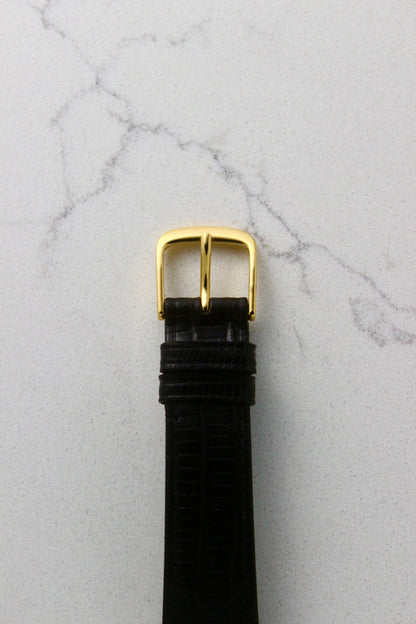 Close-up of a black lizard leather watch strap with a gold-tone buckle, photographed on a marble surface. The strap features two matching leather keepers for a secure fit.