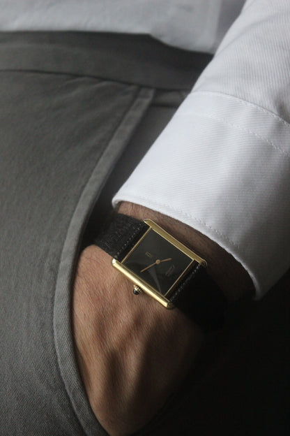 A Must de Cartier Tank watch with a yellow gold-plated sterling silver case, deep black ghost dial, and gold-tone hands. The watch is paired with a black lizard strap and features a cabochon crown. 