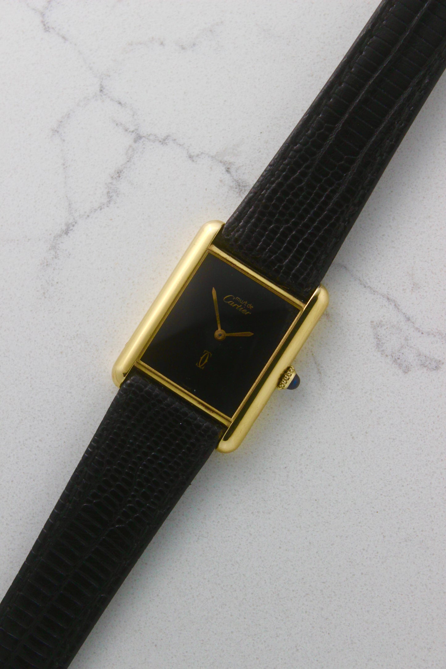 A Must de Cartier Tank watch with a yellow gold-plated sterling silver case, deep black ghost dial, and gold-tone hands. The watch is paired with a black lizard strap and features a cabochon crown. Photographed on a marble surface.