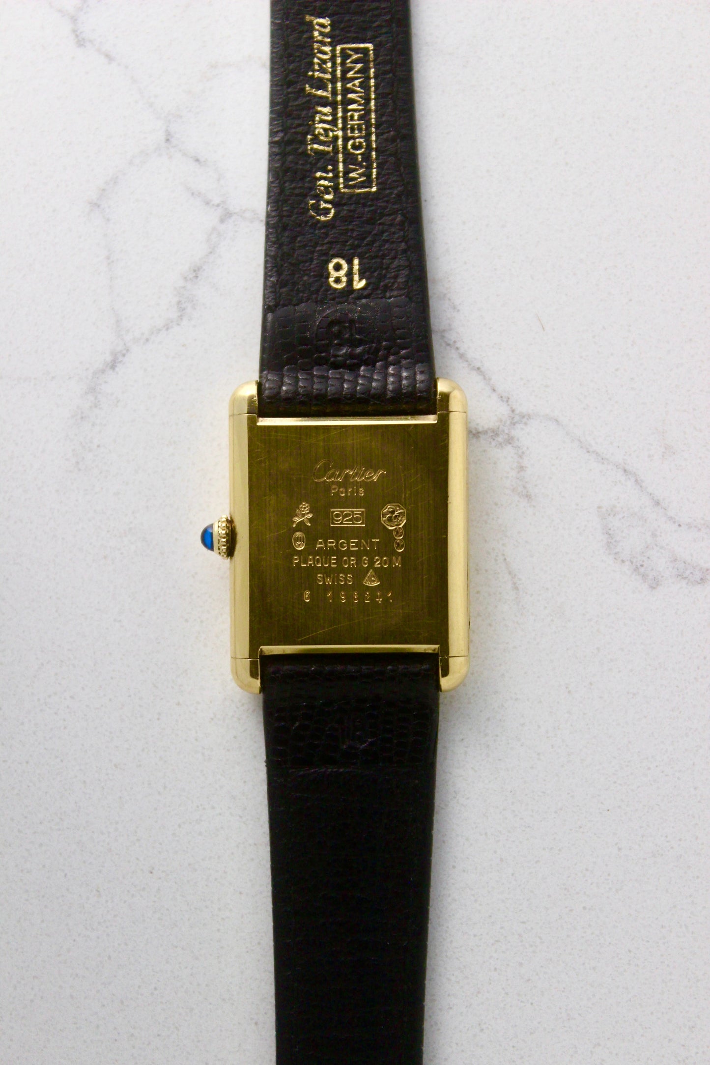 Back view of a Must de Cartier Tank watch, showing the engraved case back with 'Cartier Paris' and material details, including 925 sterling silver with gold plating. The cabochon crown is visible on the side, along with an 18 mm black lizard strap embossed with 'Gen. Lgn. Lizard W. GERMANY.' Photographed on a marble surface.