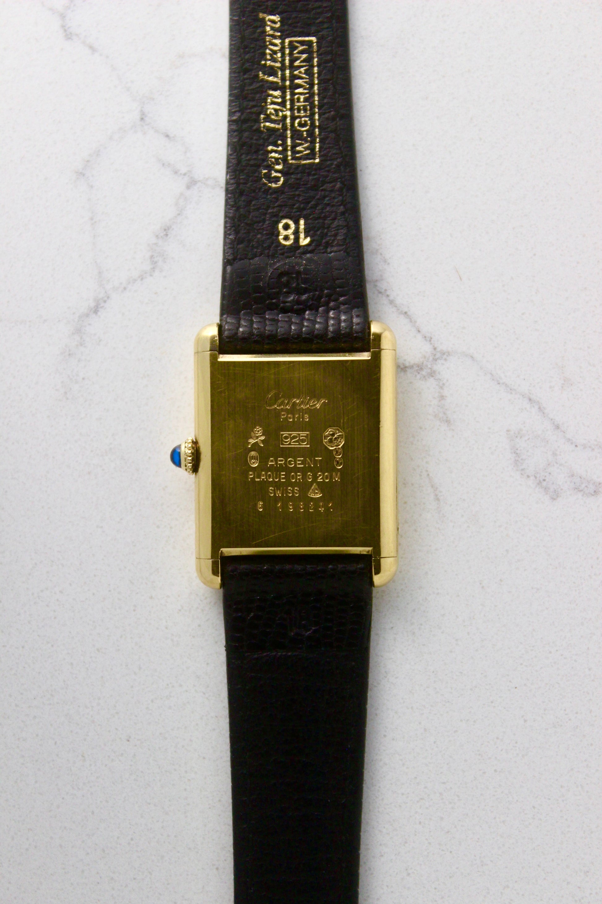 Back view of a Must de Cartier Tank watch, showing the engraved case back with 'Cartier Paris' and material details, including 925 sterling silver with gold plating. The cabochon crown is visible on the side, along with an 18 mm black lizard strap embossed with 'Gen. Lgn. Lizard W. GERMANY.' Photographed on a marble surface.
