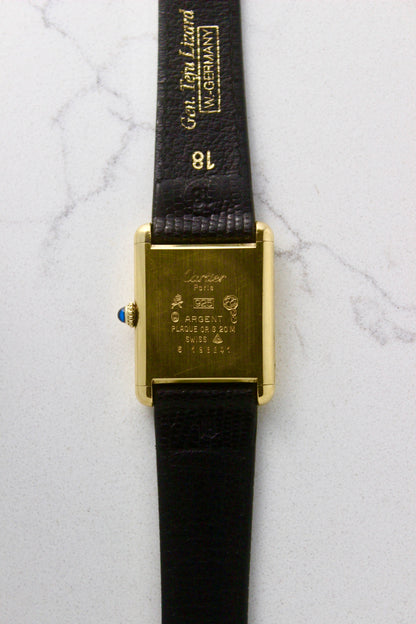 Back view of a Must de Cartier Tank watch, showing the engraved case back with 'Cartier Paris' and material details, including 925 sterling silver with gold plating. The cabochon crown is visible on the side, along with an 18 mm black lizard strap embossed with 'Gen. Lgn. Lizard W. GERMANY.' Photographed on a marble surface.