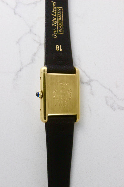 Back view of a Must de Cartier Tank watch, showing the engraved case back with 'Cartier Paris' and material details, including 925 sterling silver with gold plating. The cabochon crown is visible on the side, along with an 18 mm black lizard strap embossed with 'Gen. Lgn. Lizard W. GERMANY.' Photographed on a marble surface.