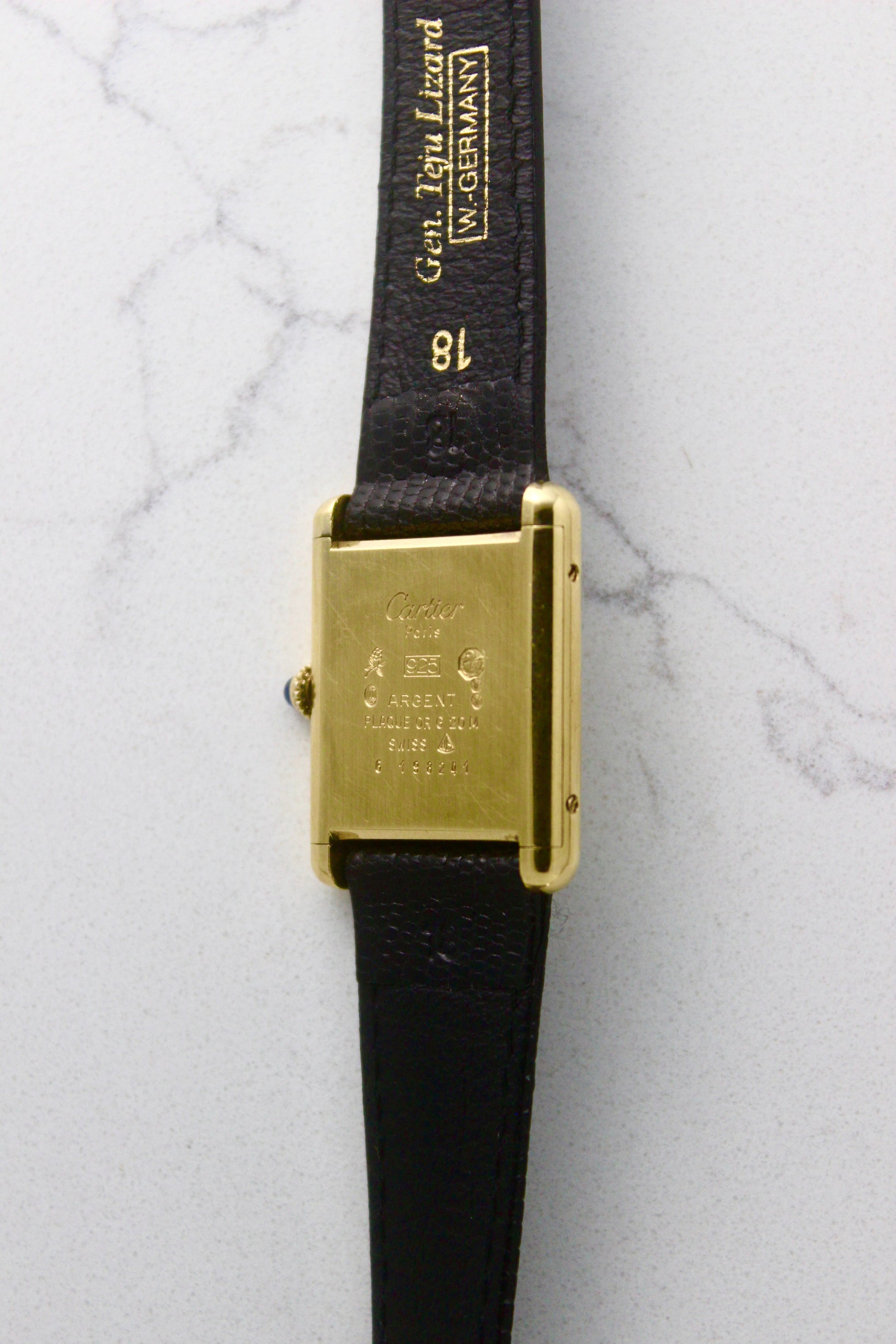 Back view of a Must de Cartier Tank watch, showing the engraved case back with 'Cartier Paris' and material details, including 925 sterling silver with gold plating. The cabochon crown is visible on the side, along with an 18 mm black lizard strap embossed with 'Gen. Lgn. Lizard W. GERMANY.' Photographed on a marble surface.