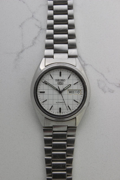 A vintage Seiko 5 automatic wristwatch (Reference Number: 7S26-3040) with a stainless steel case and bracelet. The watch features a 36mm white dial with a grid-painted design and a day-date function at the 3 o’clock position. The stainless steel bezel is fixed, complementing the original bracelet, which fits up to a 7-inch wrist. This Japan-exclusive mid-1980s model is in good condition, showing signs of wear on the bracelet, buckle, and crystal.